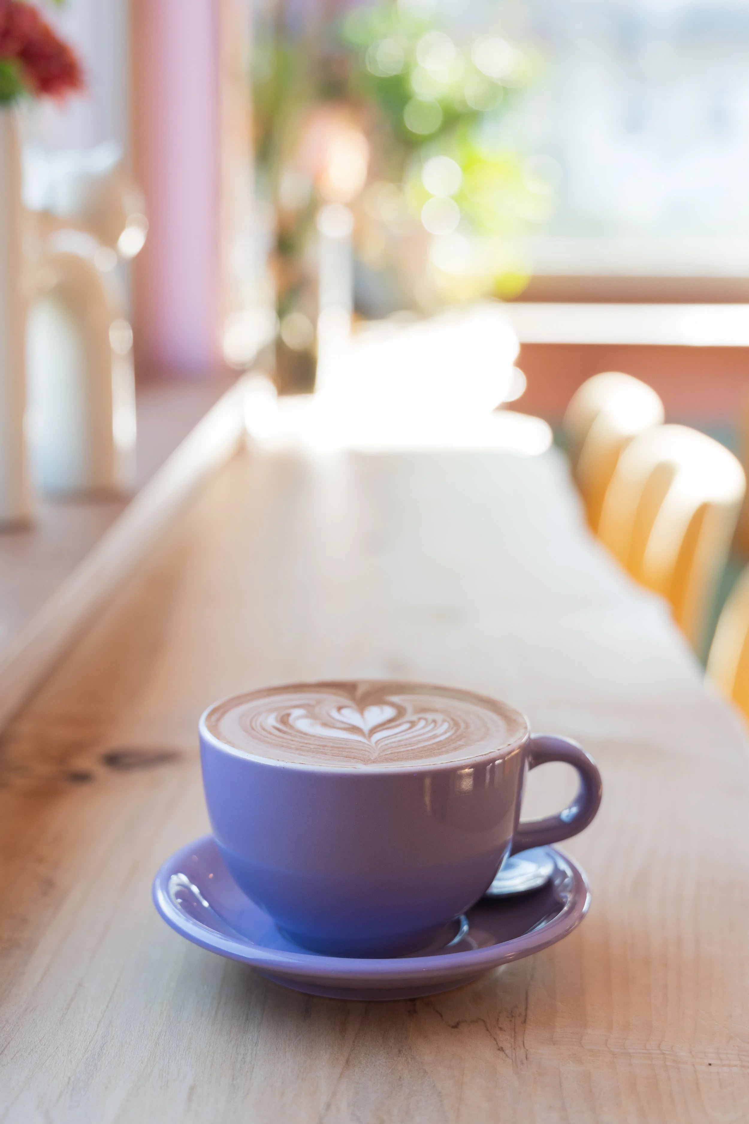 A purple coffee cup with a saucer and a spoon, containing a latte with foam art, sitting on a wooden counter in a cozy café with sunlight streaming in.