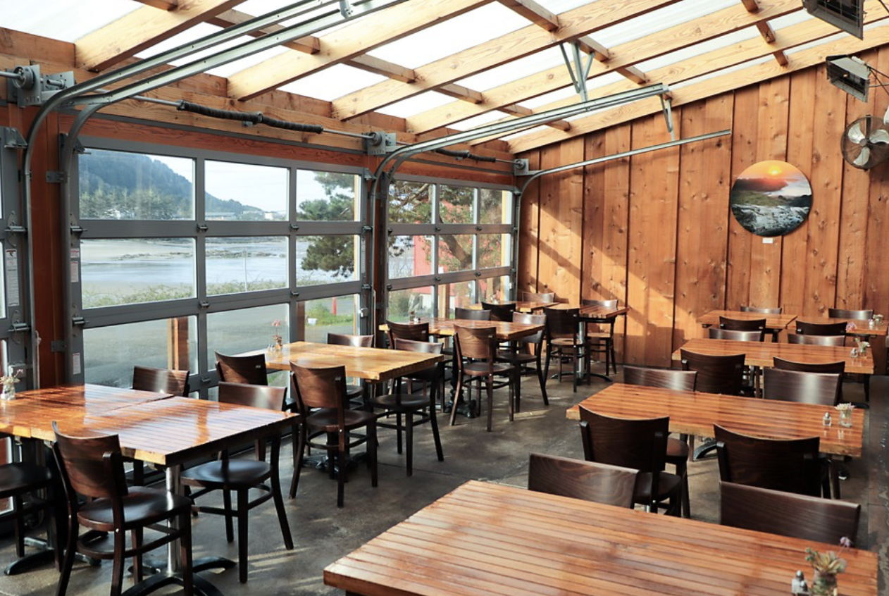 Inside a rustic restaurant with wooden walls and tables, large windows showing a river and mountains outside, and a round landscape painting on the wall.