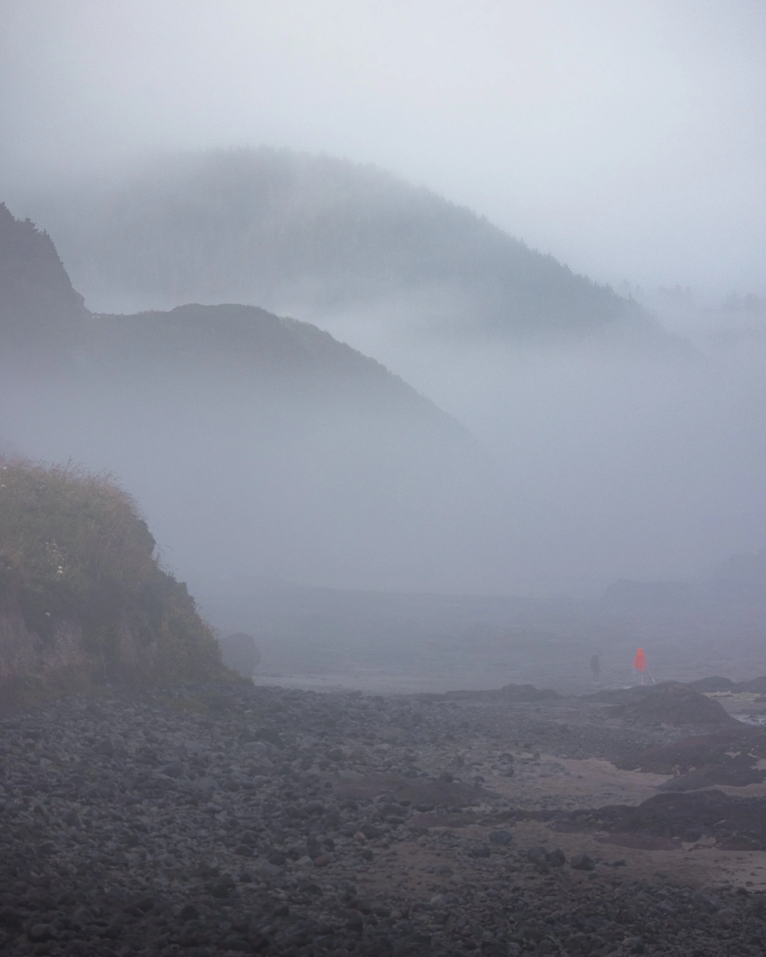 A foggy coastal landscape with mist-covered mountains in the background, rocky shoreline in the foreground, and two people walking along the beach, one in an orange jacket.