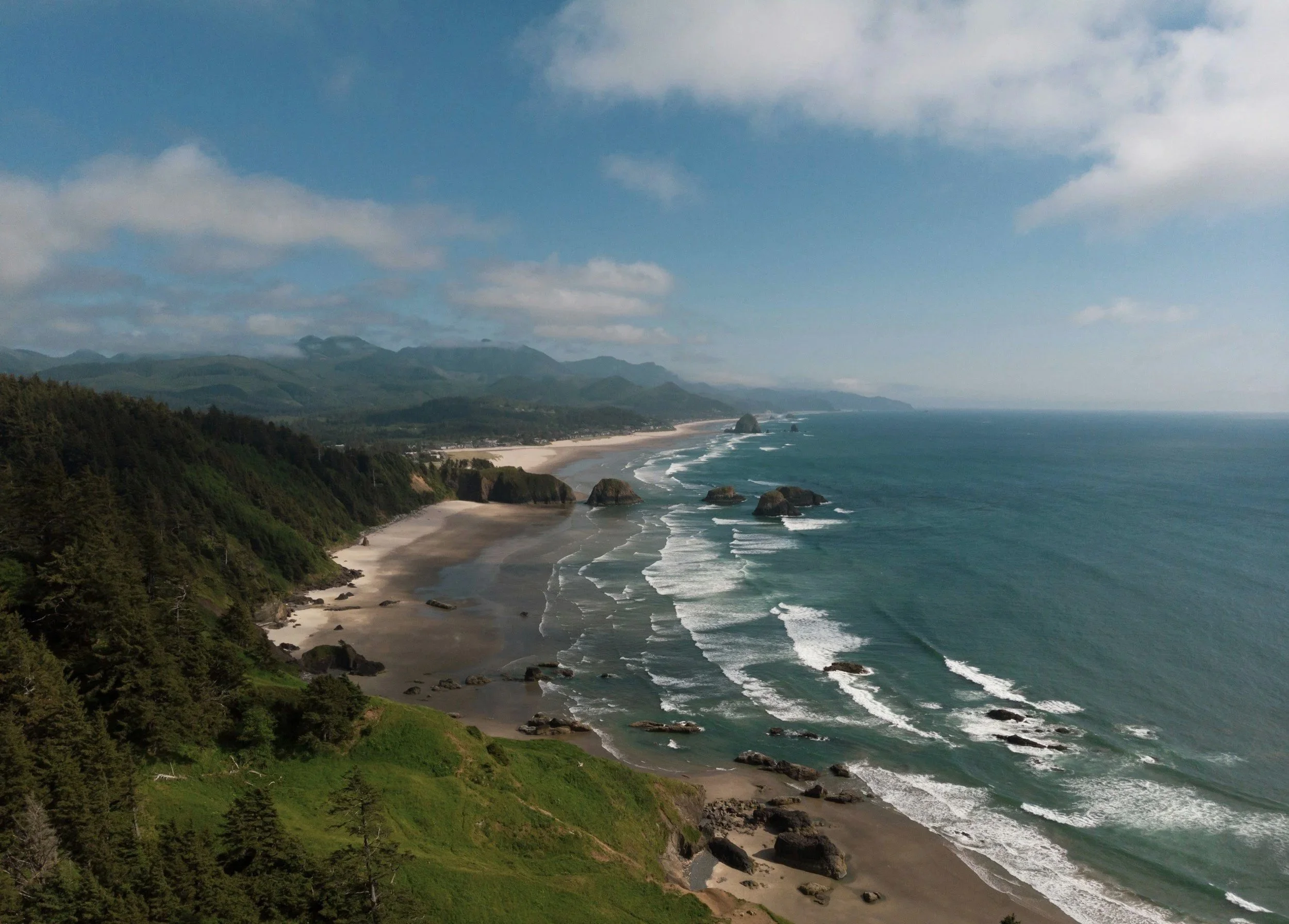 Coastal landscape with forested cliffs, sandy beaches, and large rocks in the ocean under partly cloudy sky.
