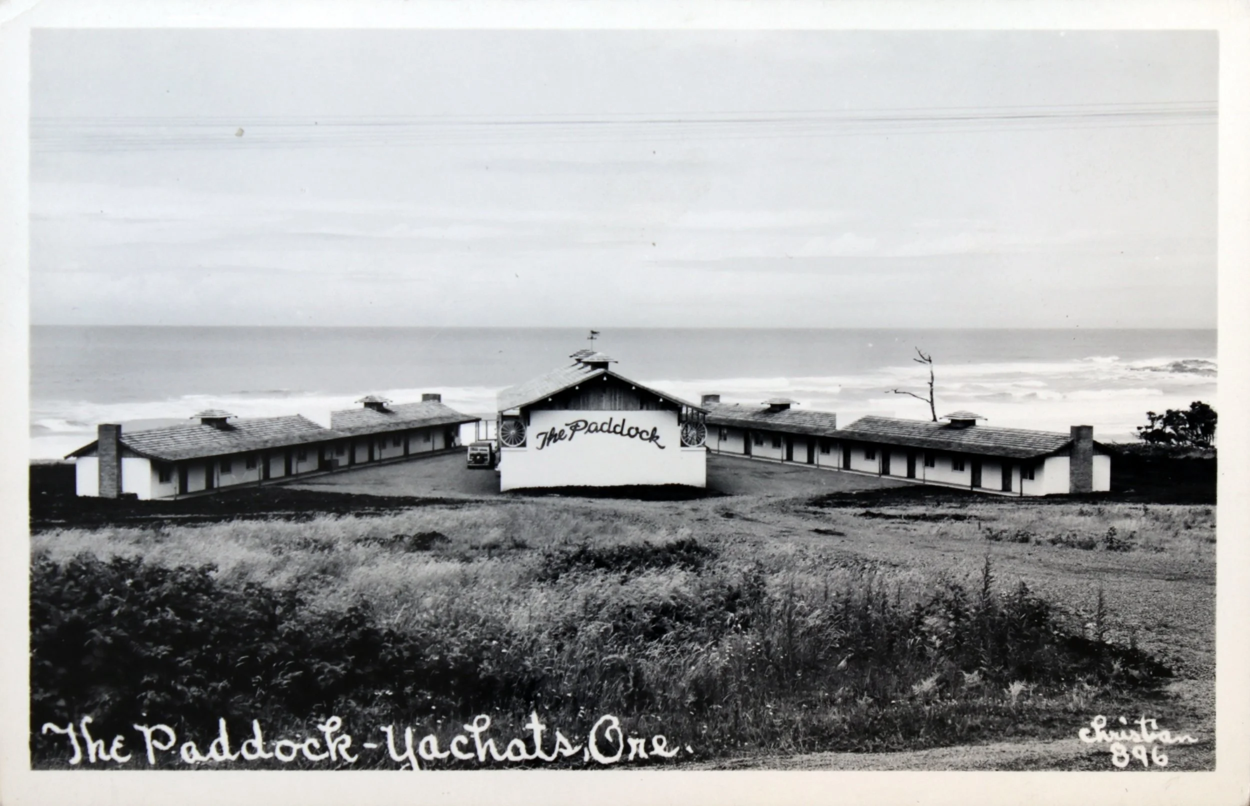 Black and white photograph of a seaside building named 'The Paddock' with a central larger structure and two extended wings, situated on a grassy hill overlooking the ocean, with a few trees and power lines in the background, and handwritten text at the bottom reading 'The Paddock - Yachats Ore' and 'Chita G. 896'.