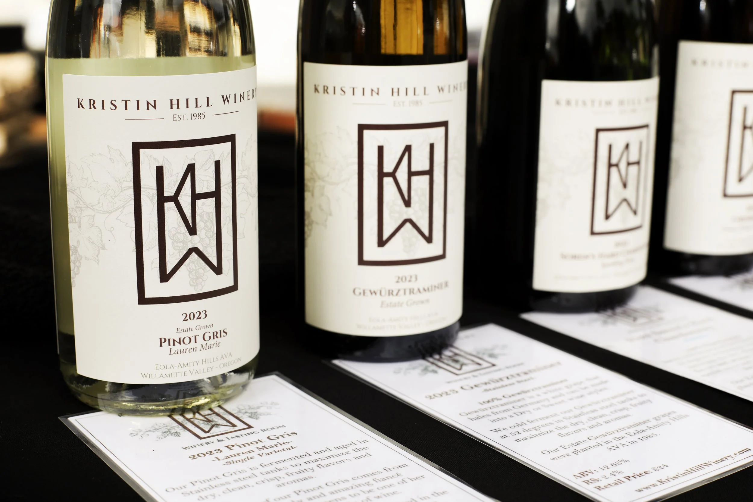 Bottles of Kristin Hill Winery wine with labels displaying the winery logo, vintage year 2023, and grape variety Pinot Gris, on a table with informational cards.