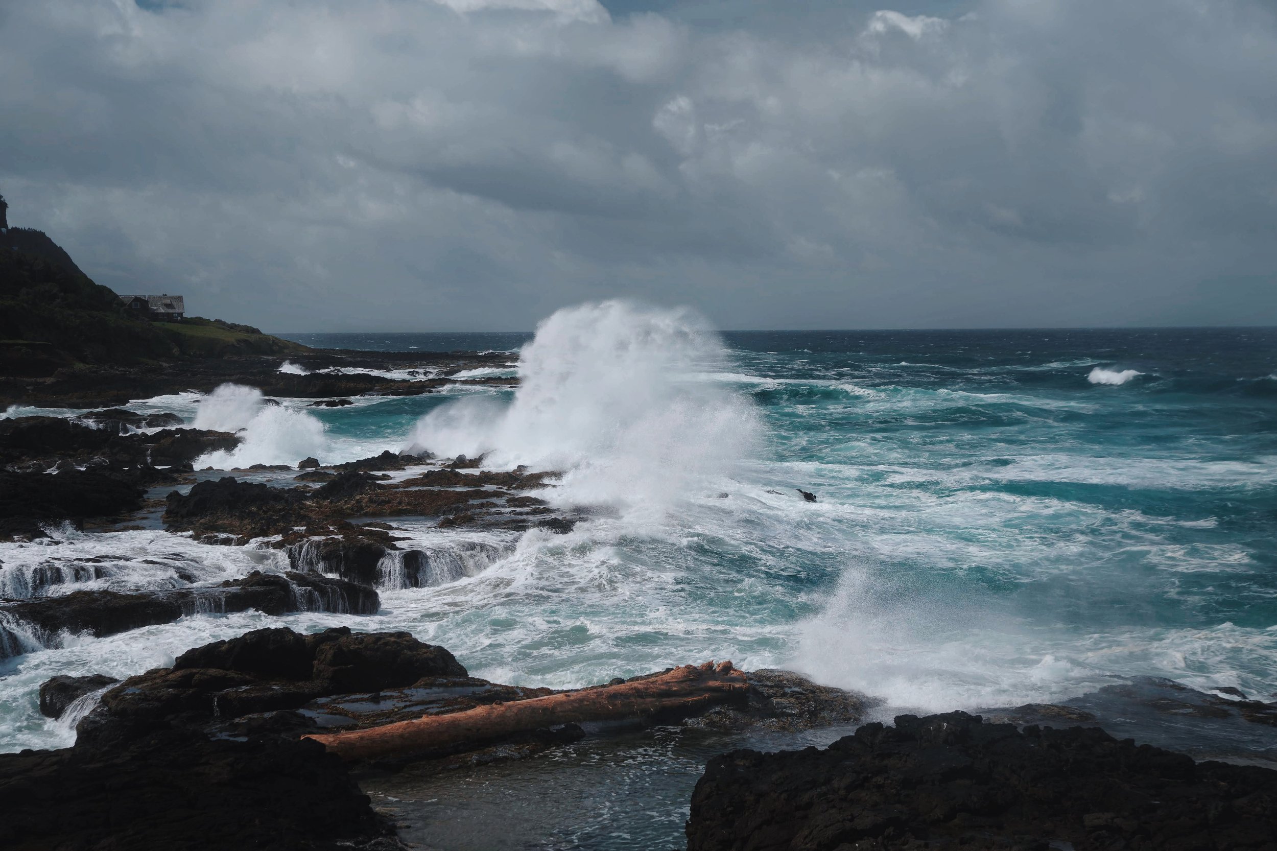 Ocean waves crashing against black rocks on a cloudy day with a house on a hill in the distance.
