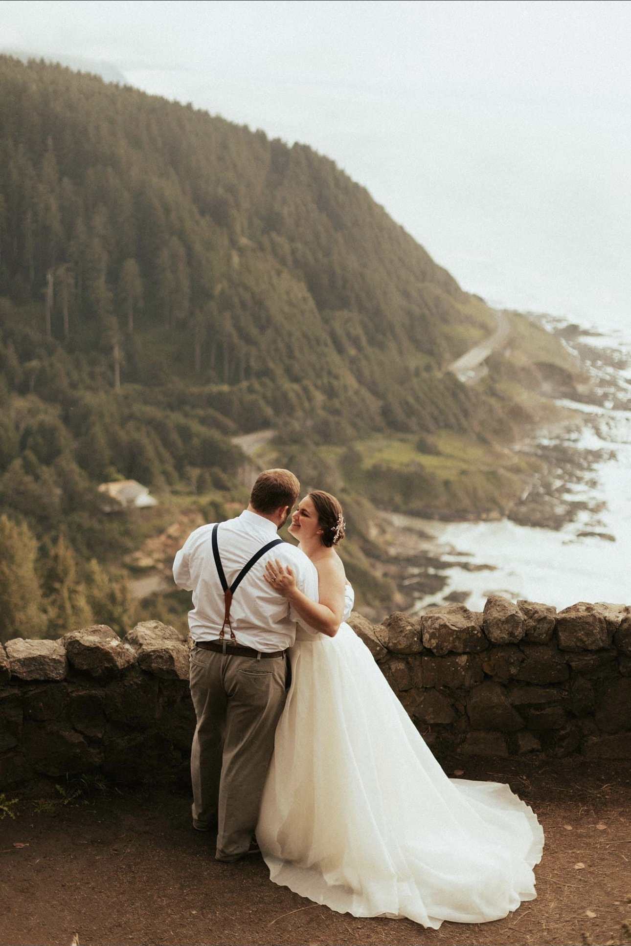 A bride and groom dancing on a rocky overlook with a scenic coastal and forested landscape in the background.