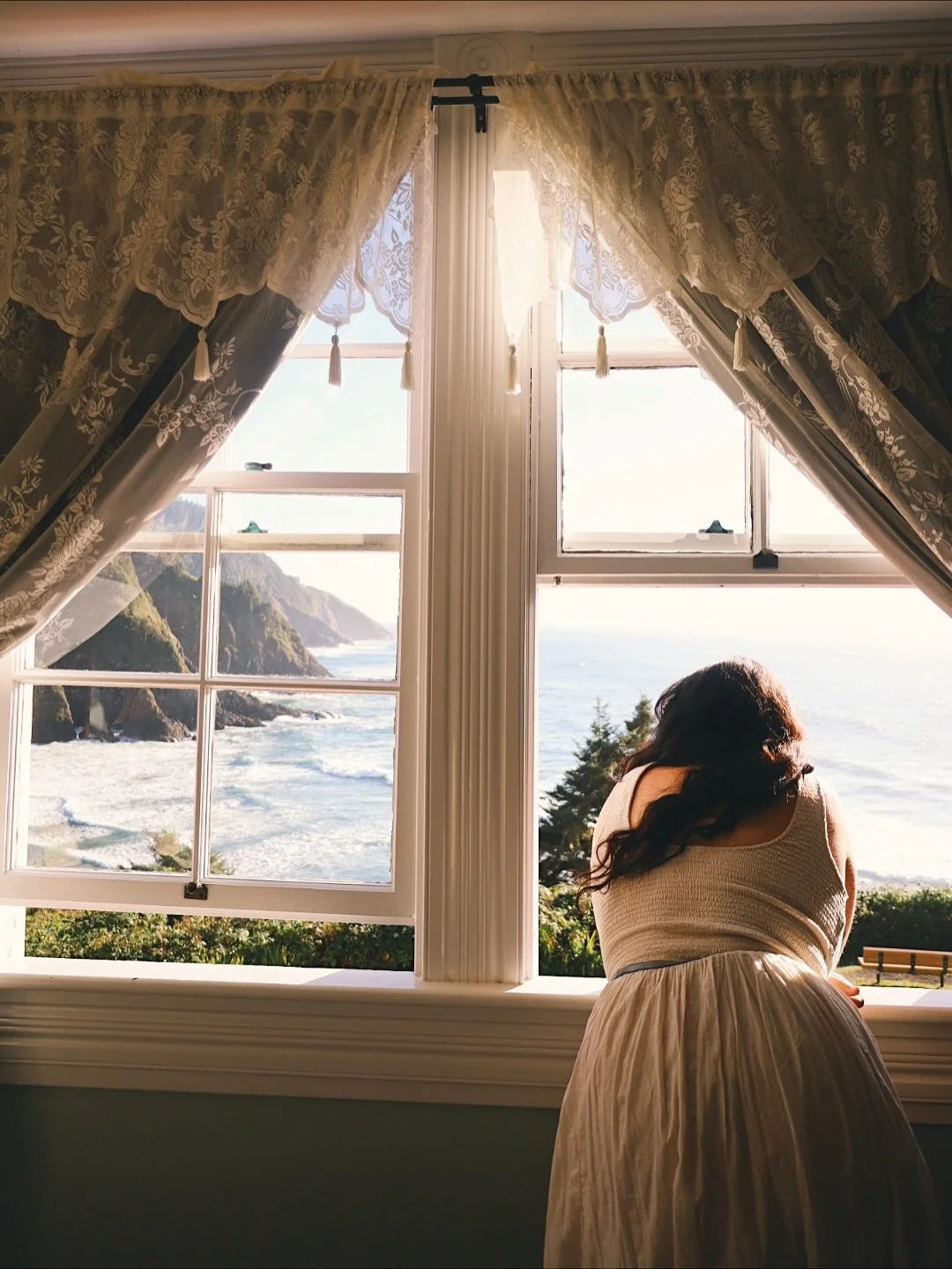A woman with dark wavy hair wearing a sleeveless white dress is leaning on a windowsill, looking out at a scenic ocean view with rocky cliffs and greenery in the distance, through open vintage lace curtains.