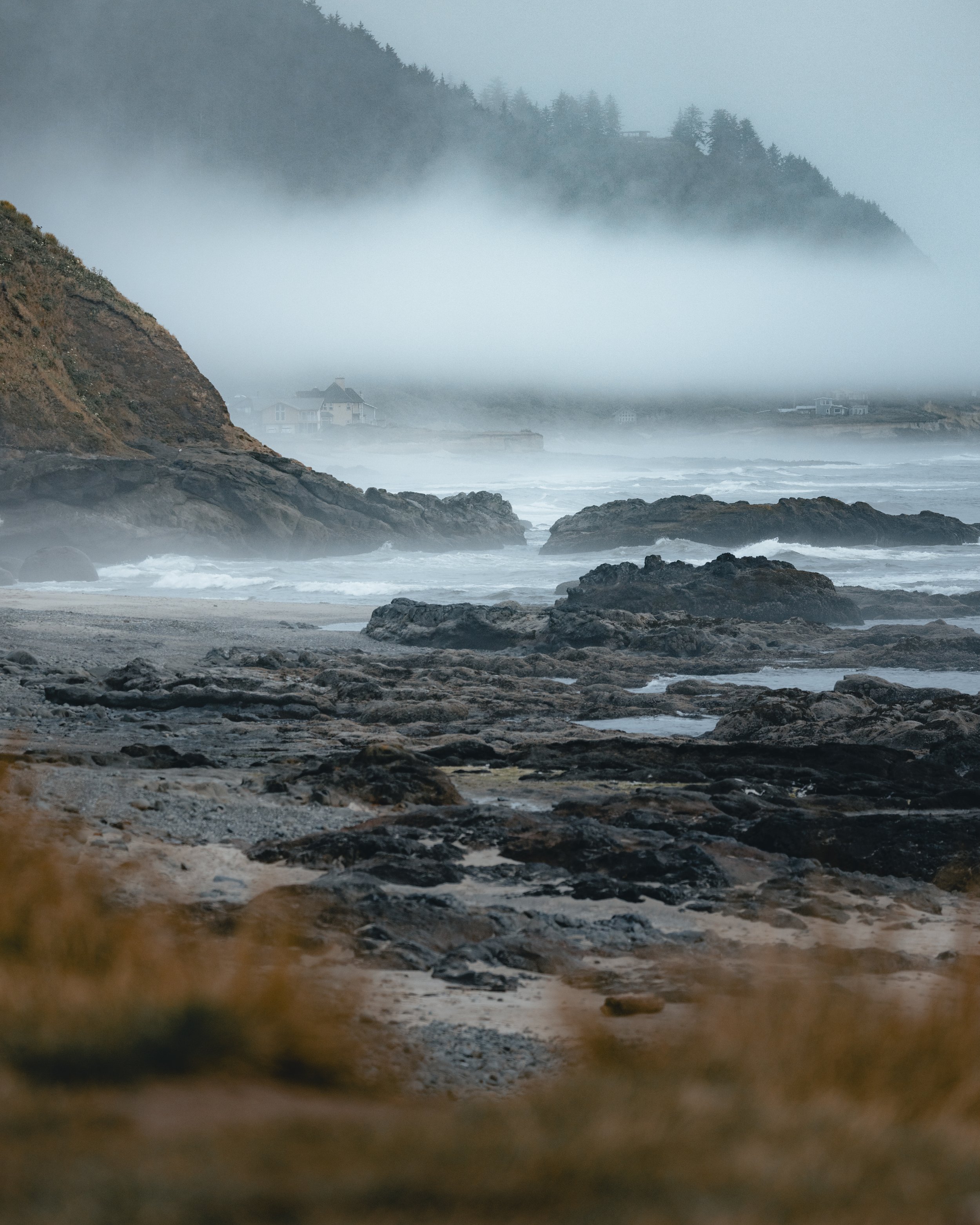 A foggy coastal scene with rocky shoreline, waves, and fog-covered hills in the background with houses visible through the mist.