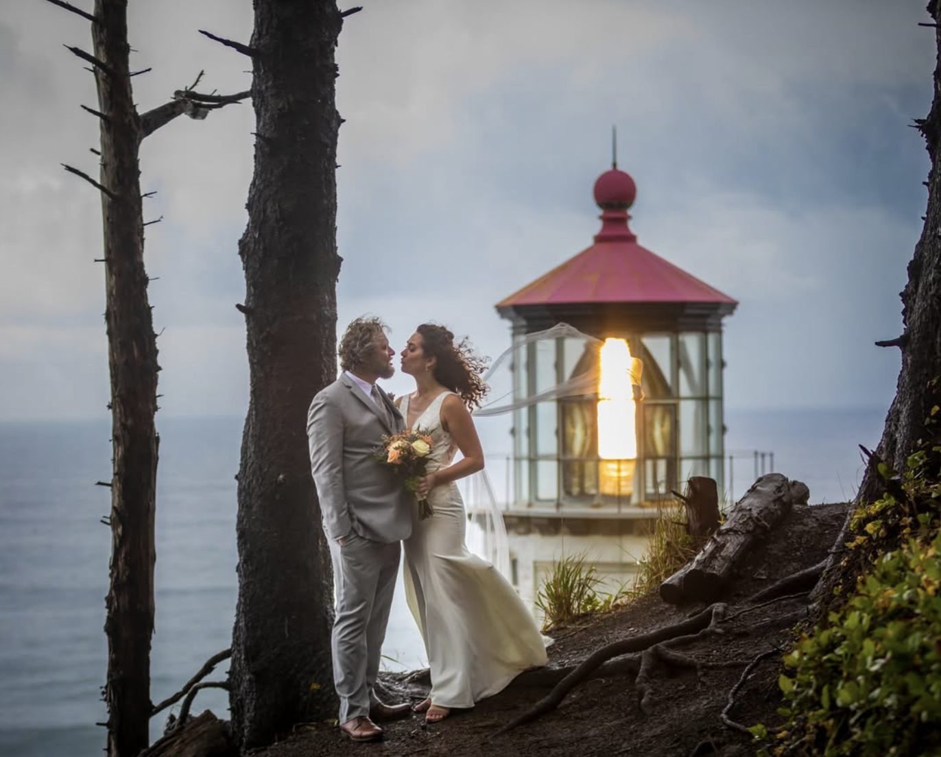 A bride and groom stand close together on a rocky shoreline at sunset, with a lighthouse in the background. The bride holds a bouquet of flowers, and they are gazing into each other's eyes.
