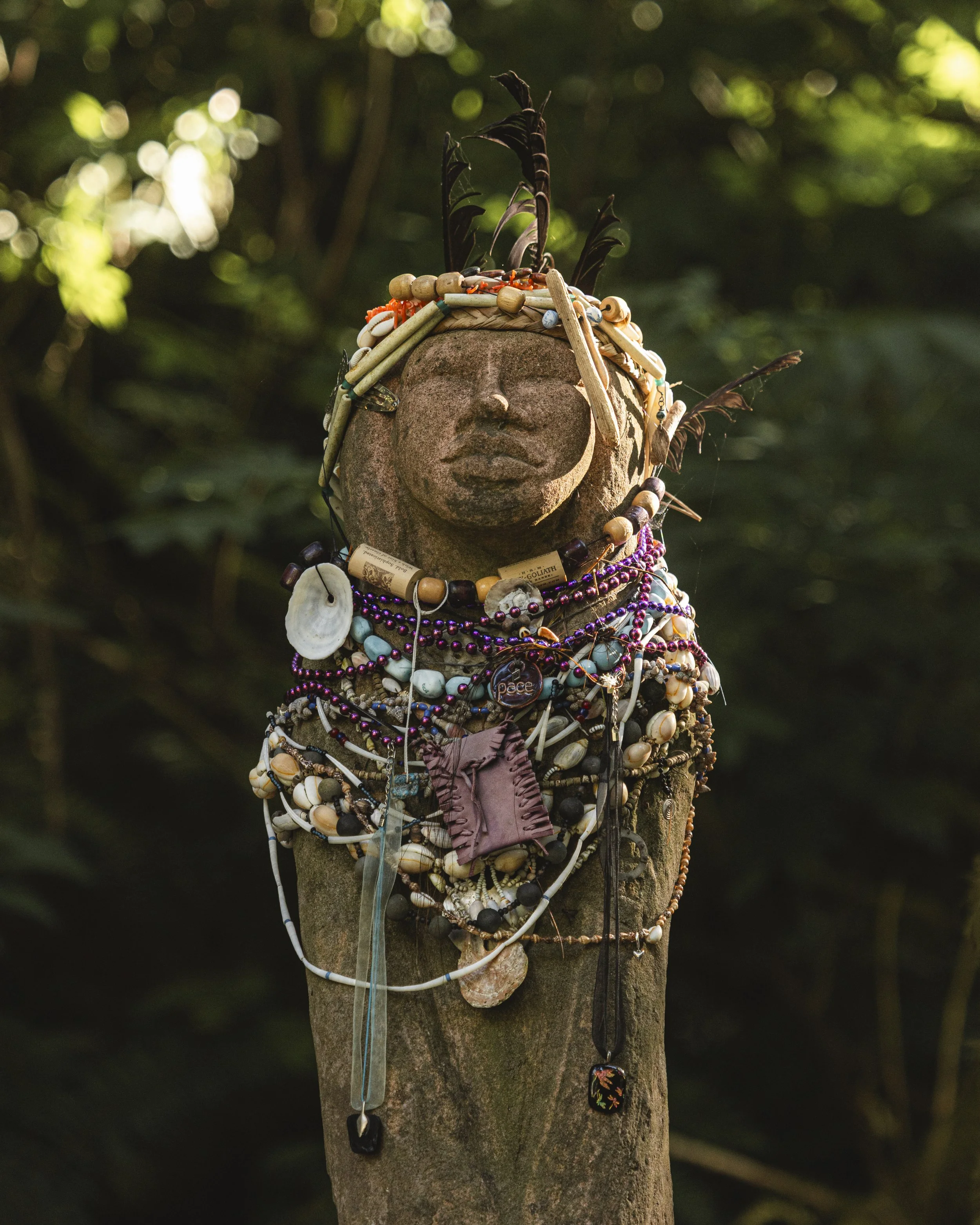 A stone sculpture of a face on a wooden post decorated with various necklaces, beads, and jewelry in a natural outdoor setting.