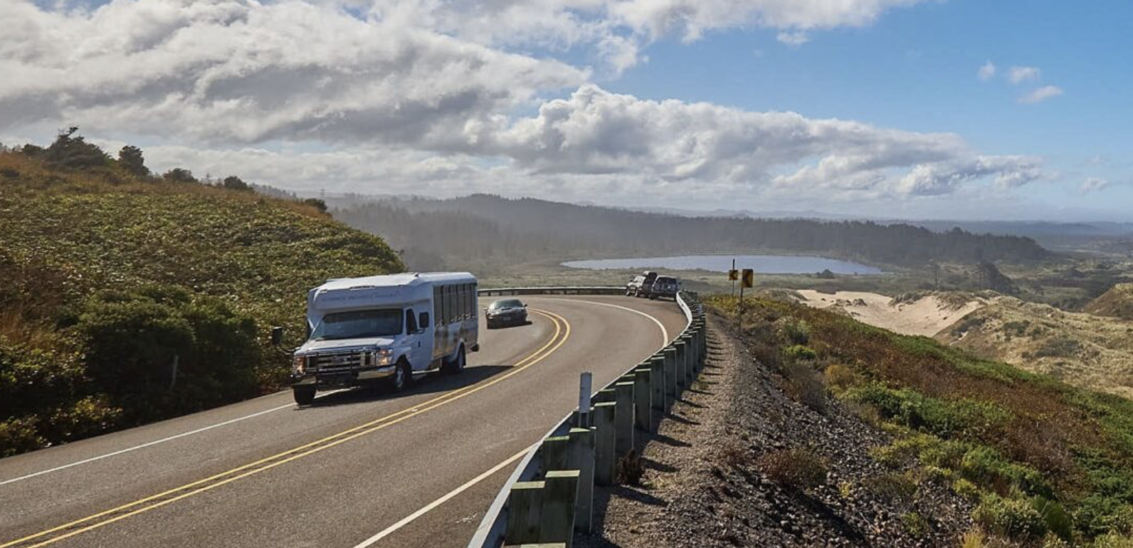A winding coastal road with vehicles, overlooking hills, a lake, and distant hills under partly cloudy skies.