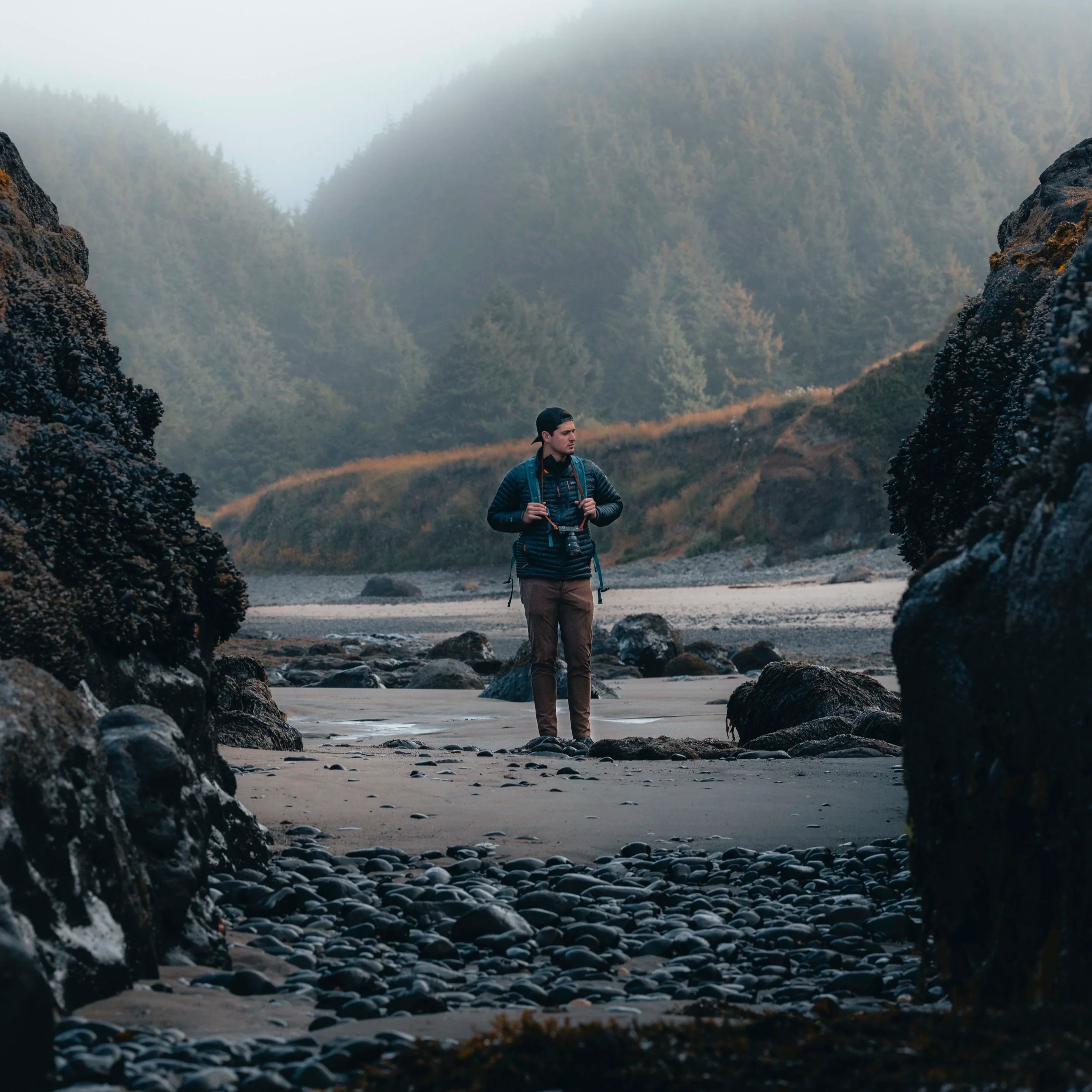 A man standing on a rocky beach, surrounded by large rocks, with forested hills in the background, wearing outdoor gear and carrying a backpack and camera.