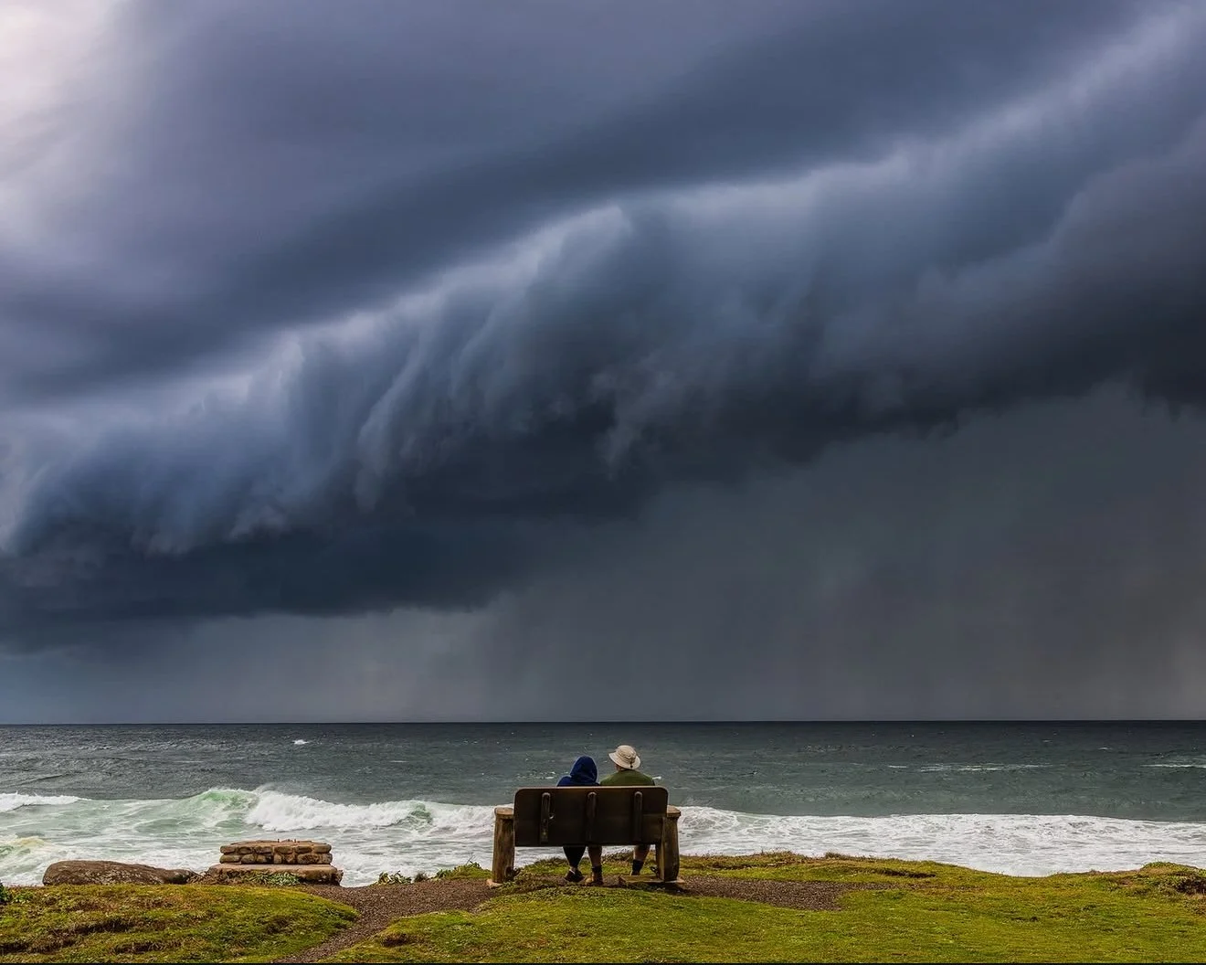 A couple sitting on a park bench facing the ocean, under a dark, stormy sky with large, ominous clouds.