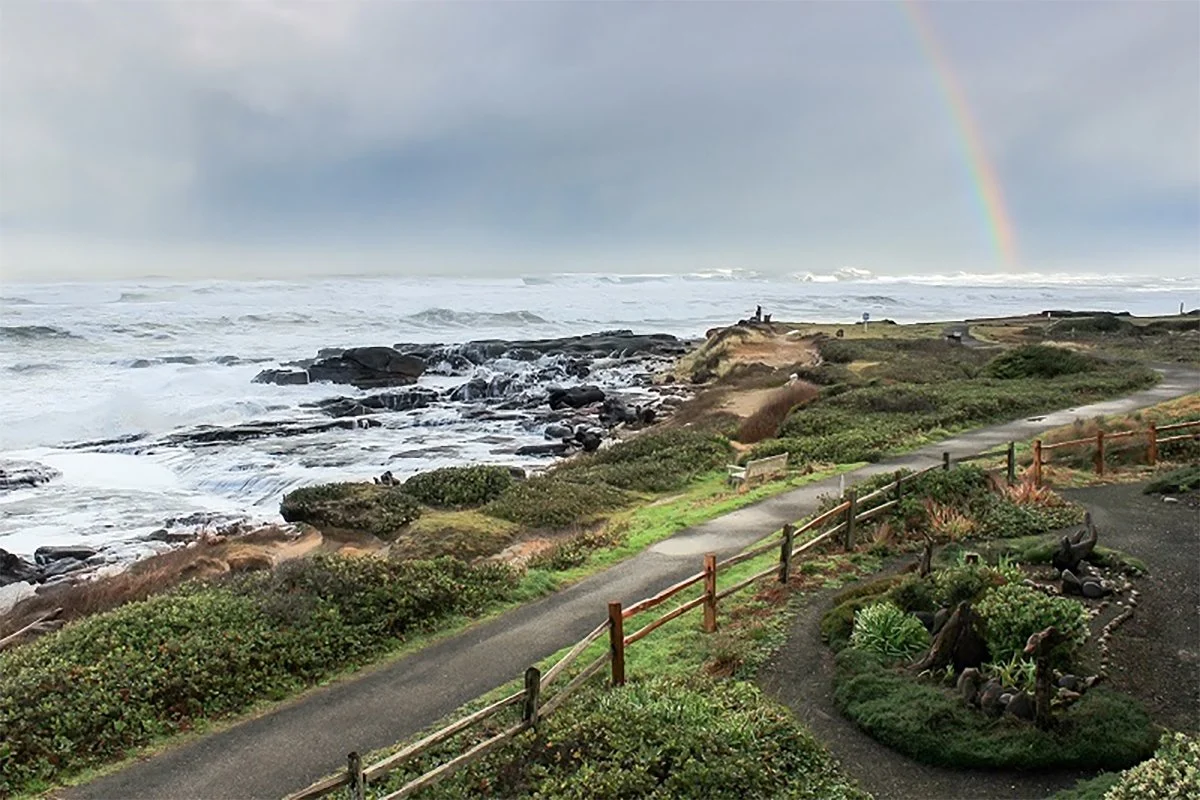 Coastal scene with a paved pathway, greenery, and a rocky shoreline with ocean waves, a rainbow visible in the cloudy sky.