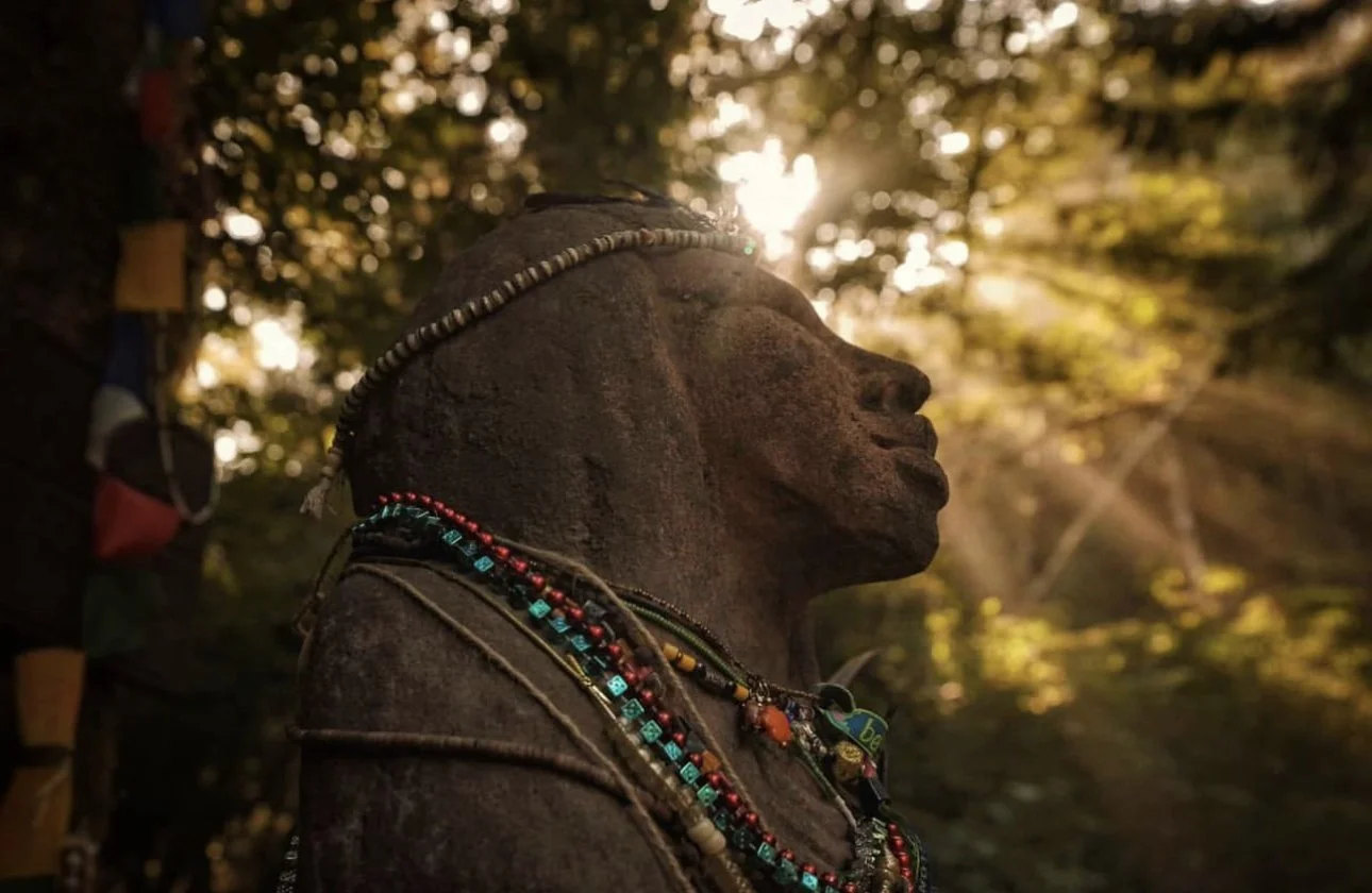 Side profile of a stone statue of a woman adorned with colorful beaded necklaces and a beaded headband, with sunlight filtering through trees in the background.
