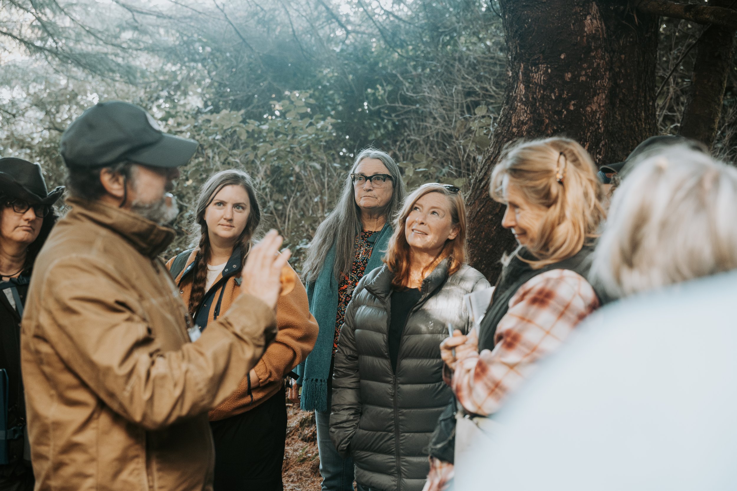 A group of people listening to a man in a brown jacket and black cap speaking outdoors in a forested area.