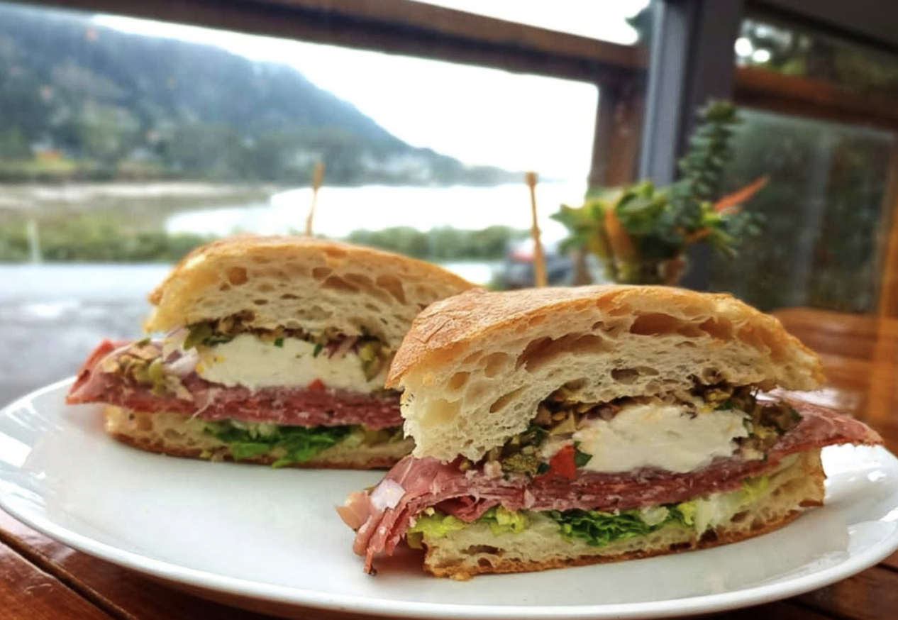 Close-up of a sandwich on a white plate with layers of meat, cheese, and lettuce, set on a wooden table with a scenic outdoor view in the background.