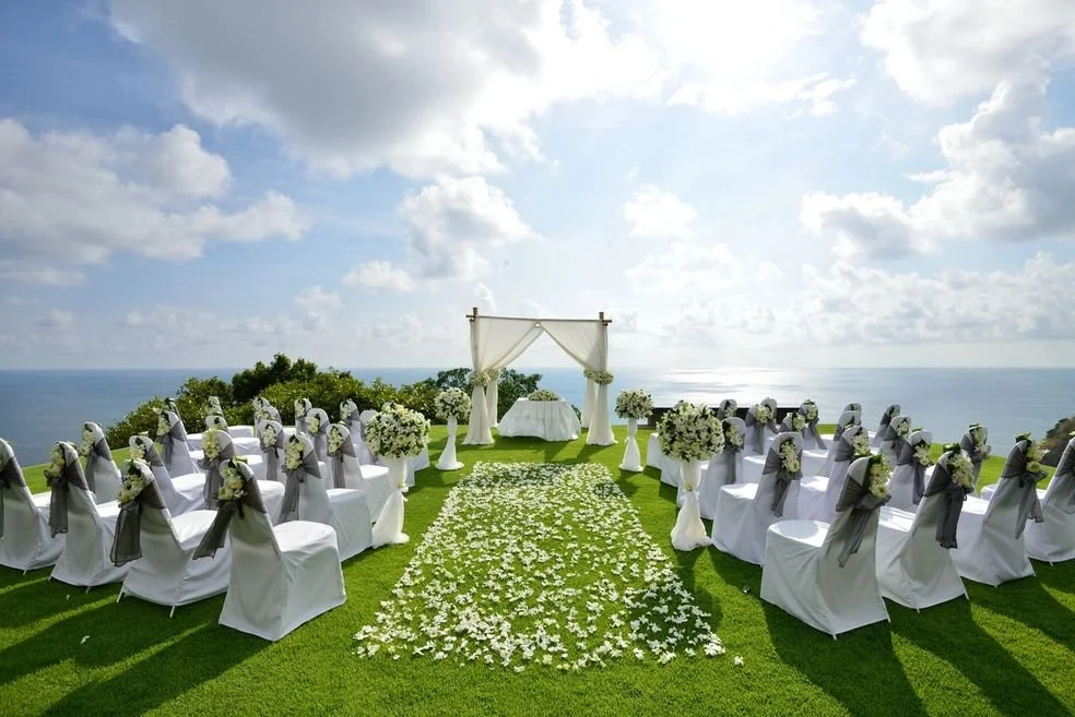 Outdoor wedding setup on a grassy lawn overlooking the ocean with white chairs, decorated with gray sashes and floral arrangements, leading to a white arch with sheer curtains.
