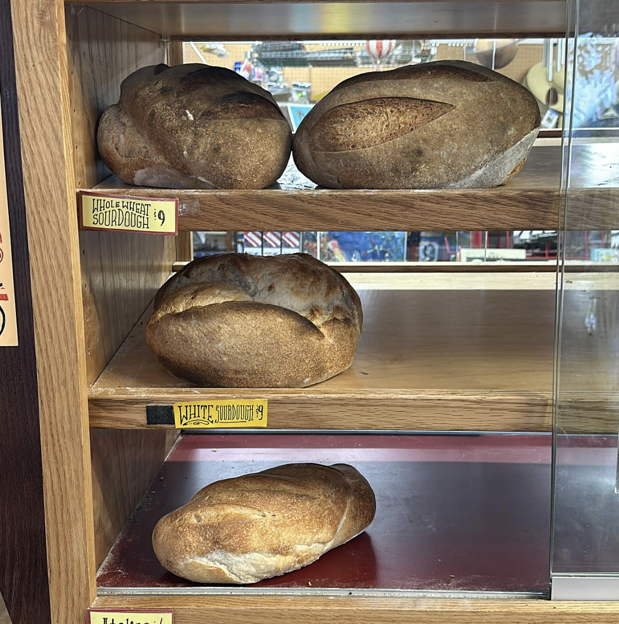 Three loaves of bread on wooden shelves in a bakery display case, labeled as whole wheat sourdough, white sourdough, and an unidentified bread.