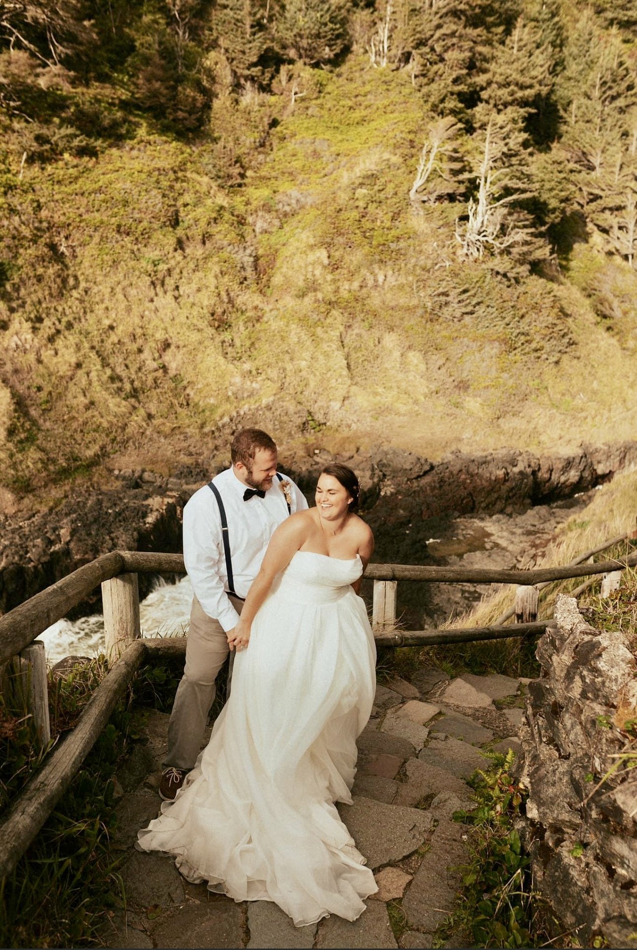 A couple dressed in wedding attire standing on a stone pathway with a wooden railing on a cliffside overlooking a waterfall and lush green mountains at sunset.