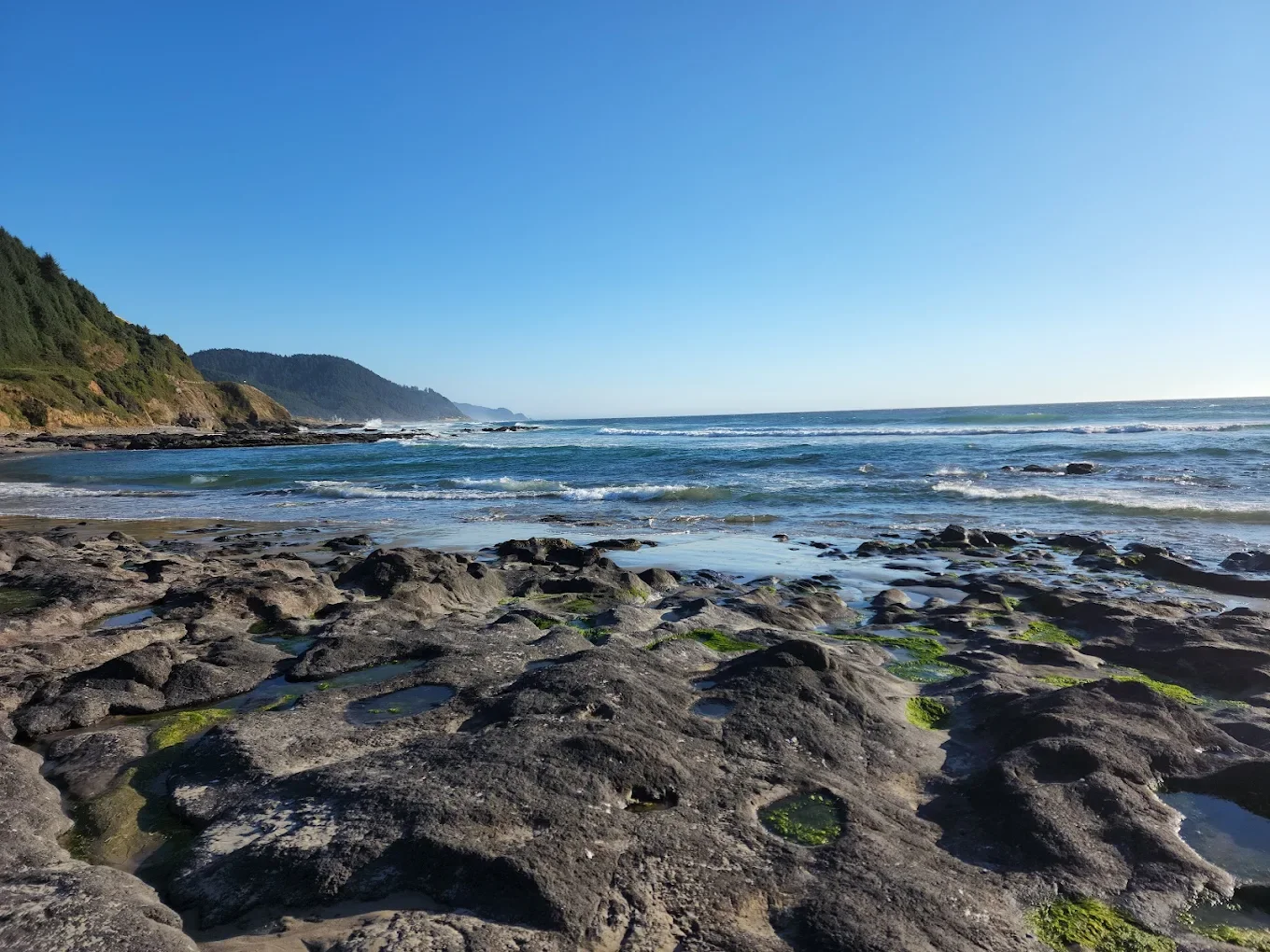 Rocky shoreline with green moss, ocean waves, and a distant hillside under a clear blue sky
