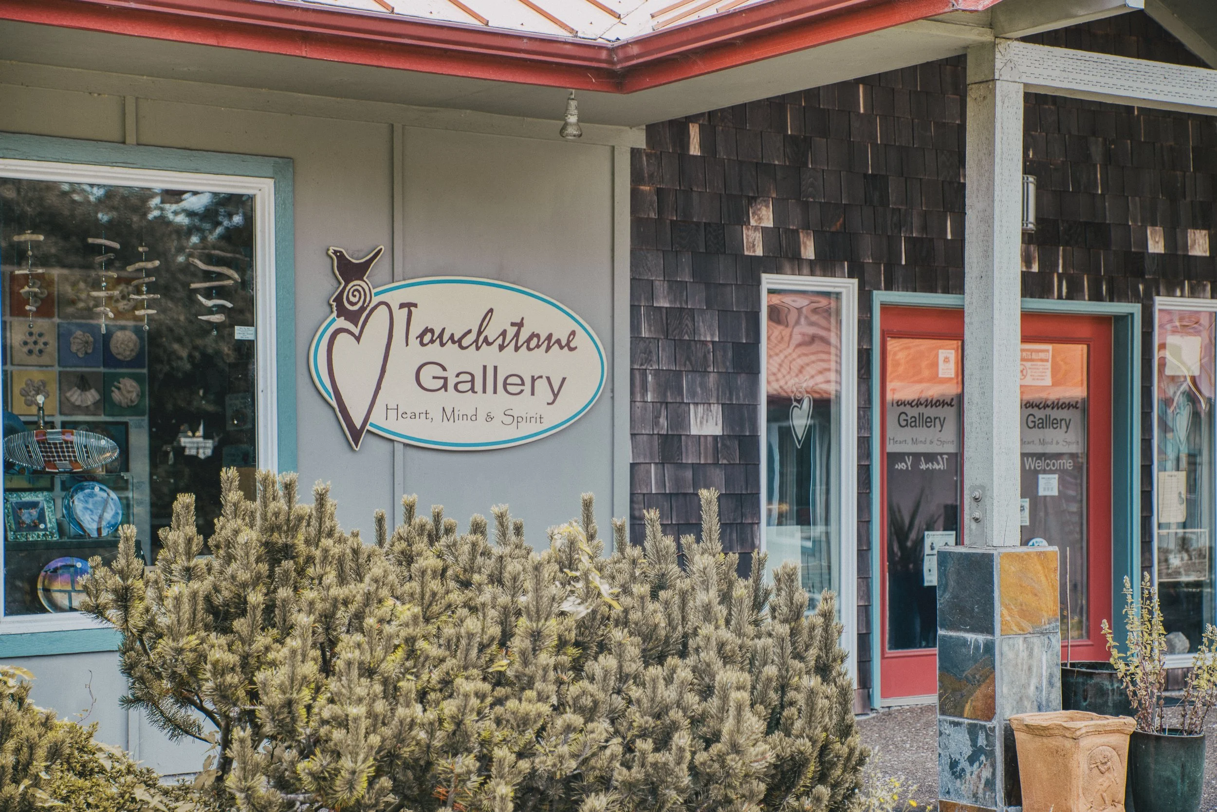 Exterior of a store called Touchtone Gallery with a sign reading "Heart, Mind & Spirit." The entrance has red doors, and there is a window display with art and souvenirs. There are potted plants and bushes outside.