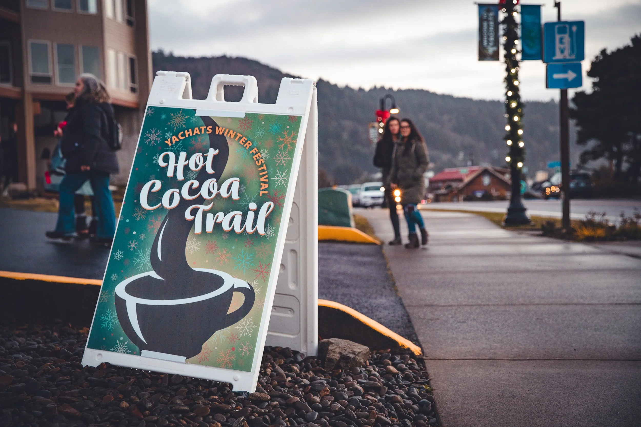 Sign advertising Hot Cocoa Trail at Yachats Winter Festival with people walking on the sidewalk in a mountain town at dusk.