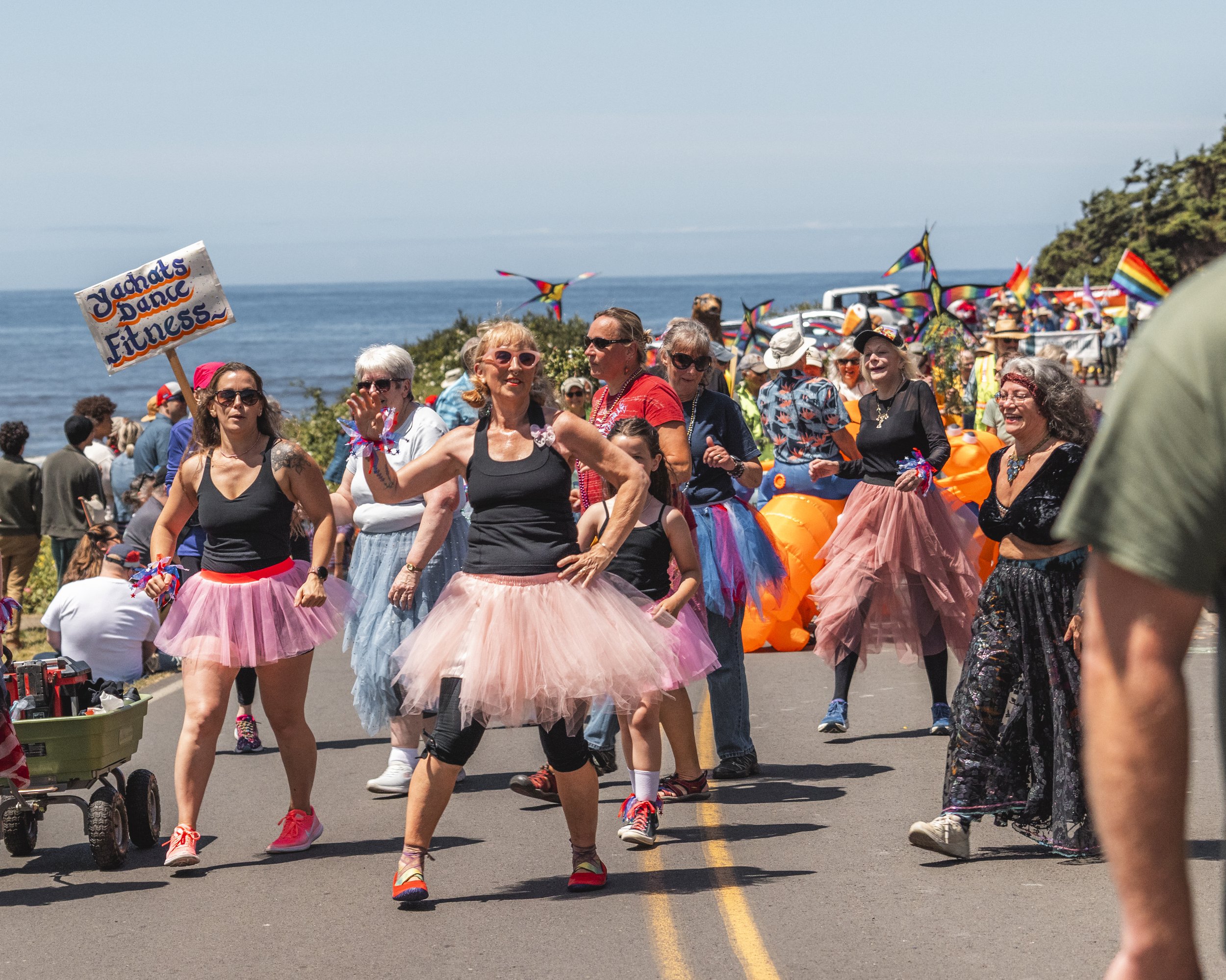 People dancing in colorful tutus at a parade near the coast, with rainbow flags and decorations in the background.