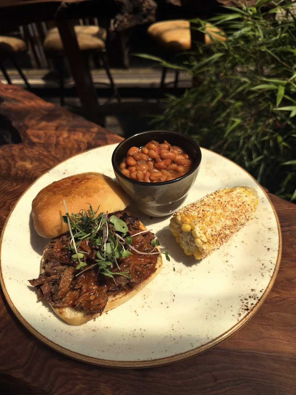 Plate with a bun sandwich topped with shredded beef and microgreens, a baked bread roll, a bowl of baked beans, and a piece of grilled corn on the cob topped with seasoning, on a white ceramic plate.