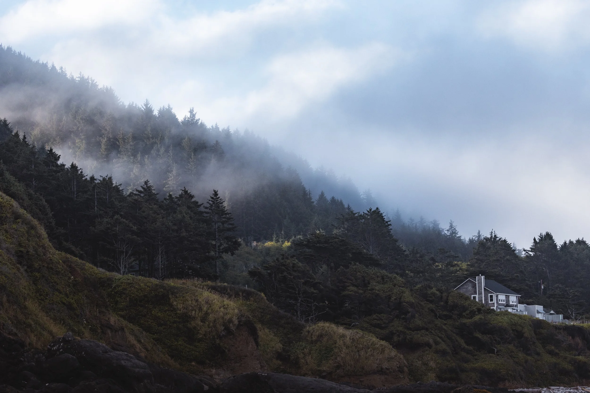 A house on a hilly, forested coastline with fog and clouds in the sky