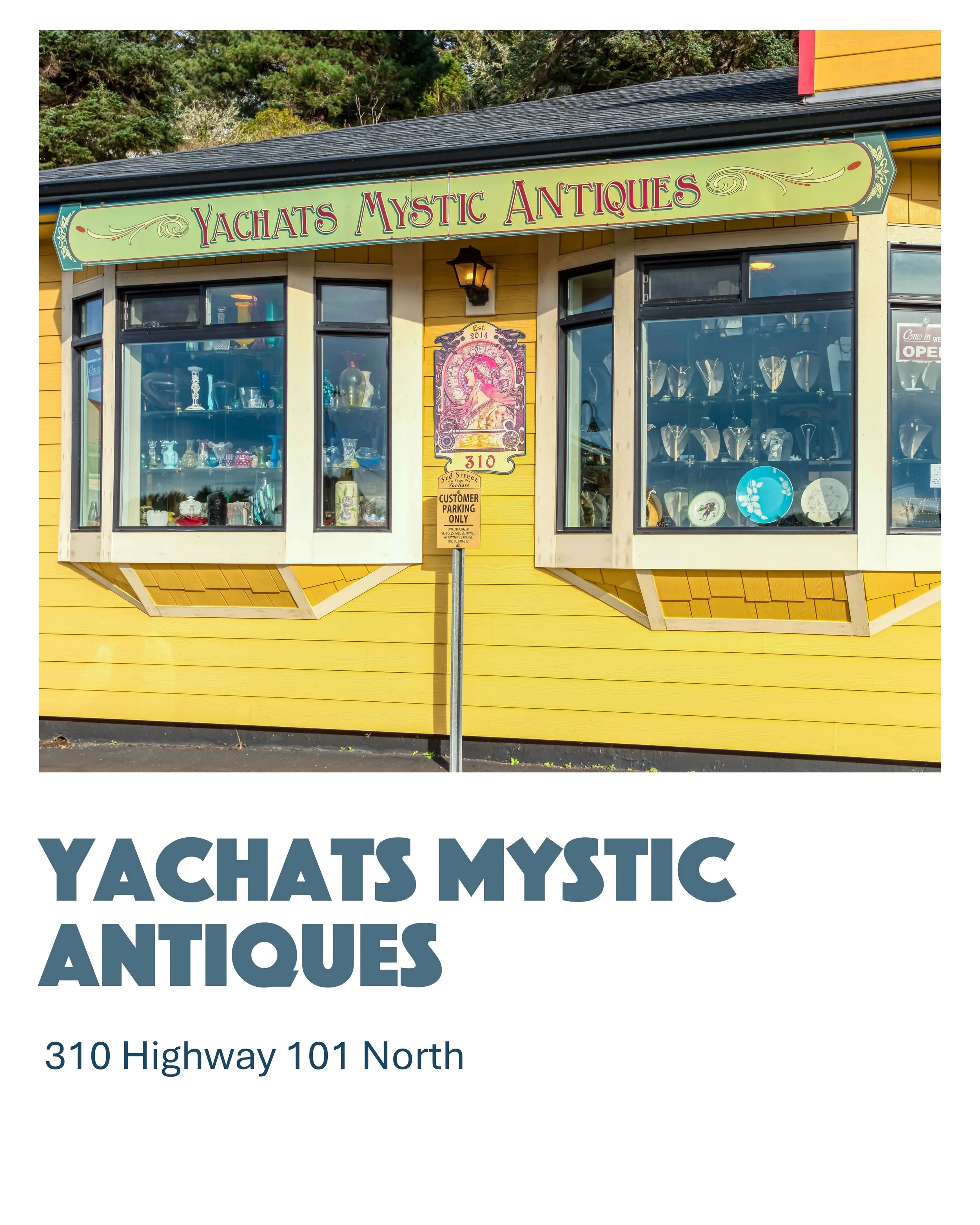 A yellow building with large display windows showcasing various antiques including plates, glasses, and decorative items. The store sign reads "Yachats Mystic Antiques" and is green with red and purple lettering. There is a street lamp, an address plaque, and a parking sign in front. The background features green trees.