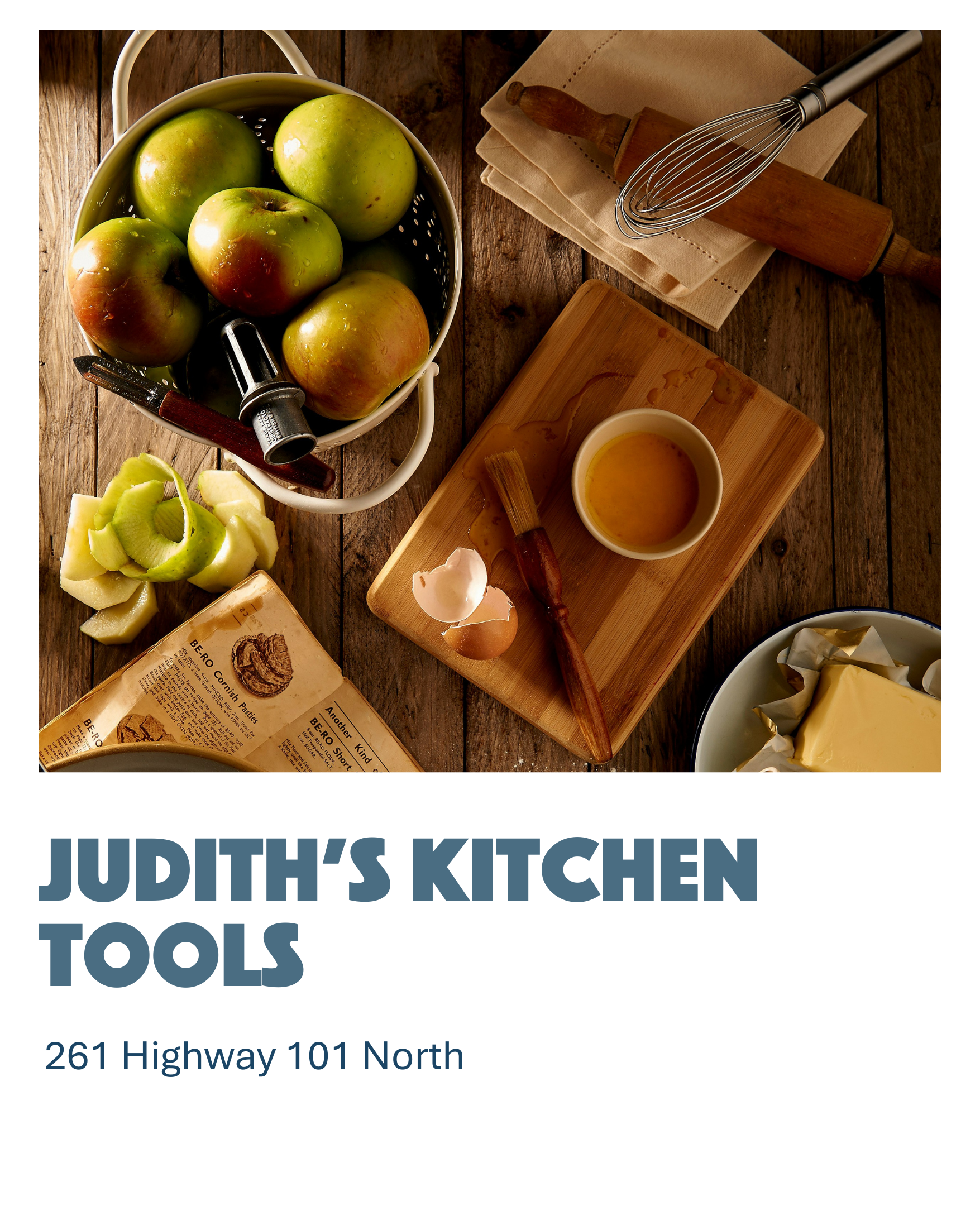 A top-down view of a kitchen countertop with apples, a lemon, a butter wrapper, a wooden rolling pin, a whisk, a small bowl of oil, a wooden cutting board, and a butter dish.