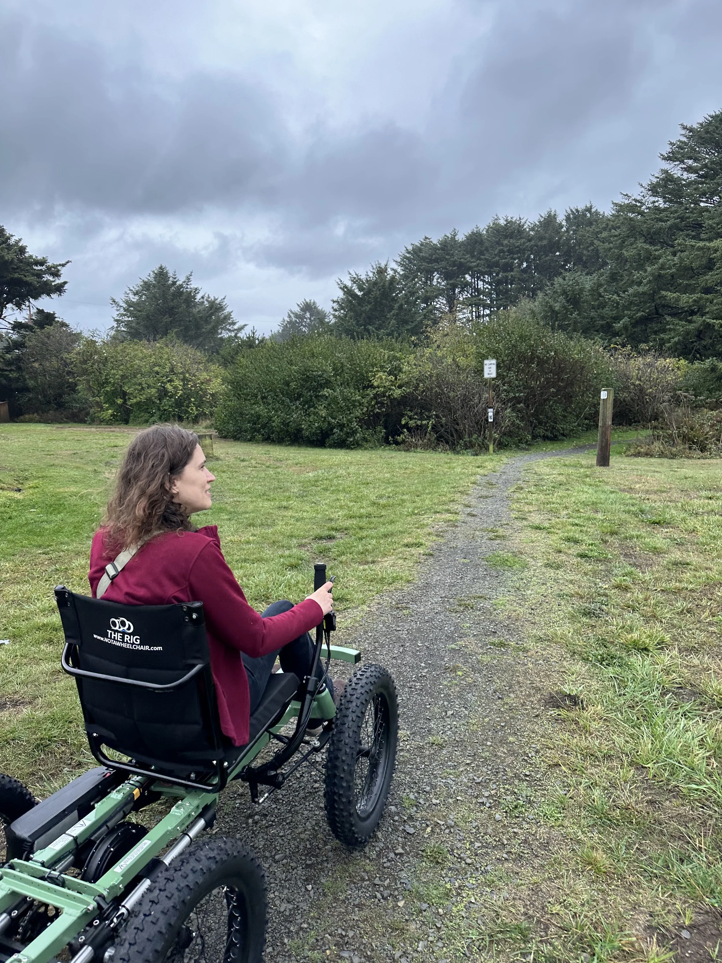 Woman in a wheelchair riding on a gravel trail through a grassy field and trees on a cloudy day.