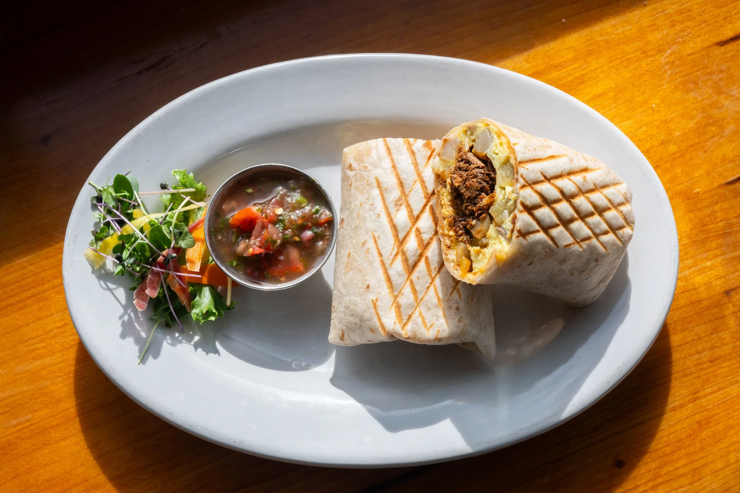 A plate with a grilled wrap cut in half, showing beef, potatoes, and other fillings inside, with a side of salad greens and salsa.