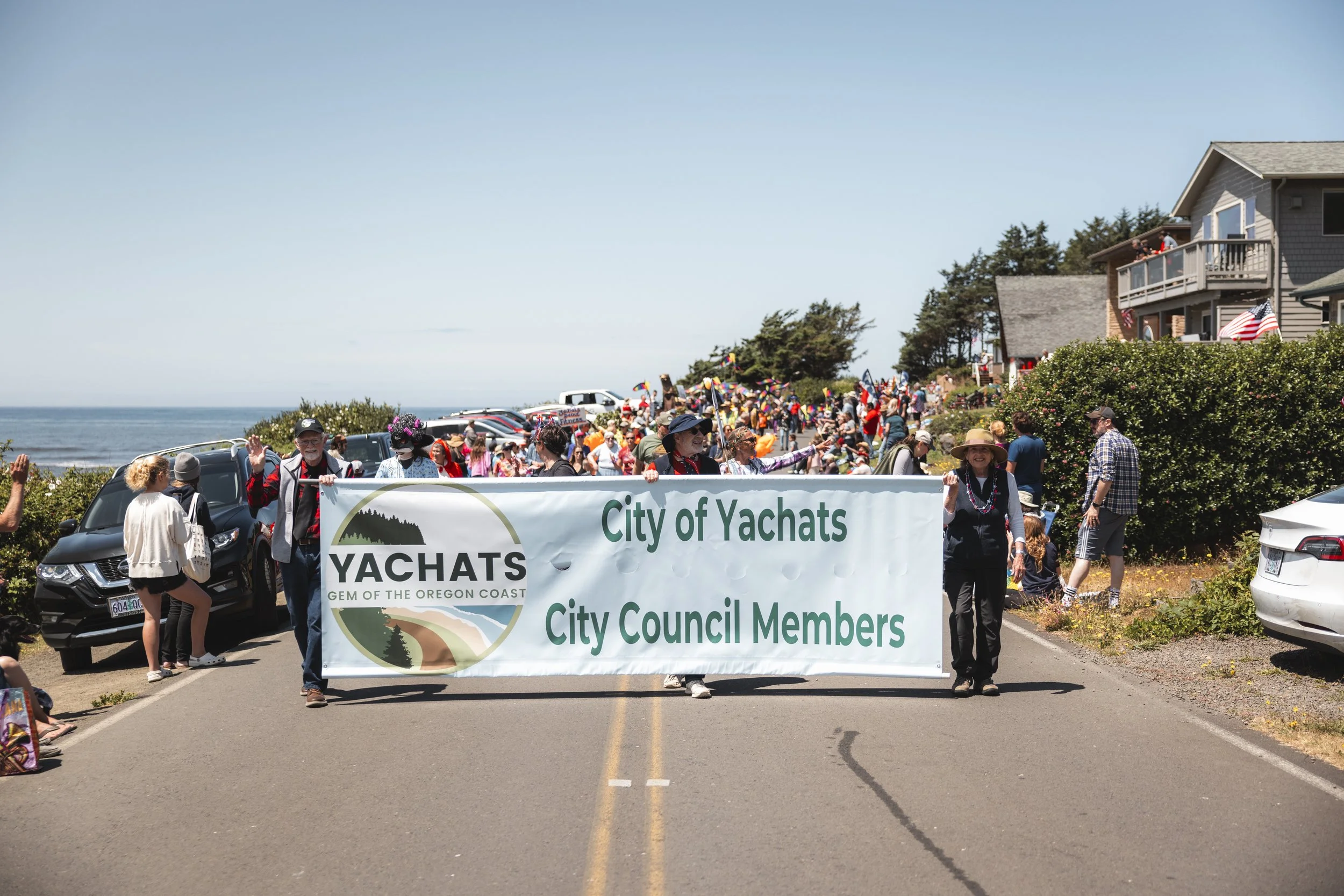 People holding a large banner that reads 'City of Yachats, City Council Members' during a parade or protest near the coast with houses and trees in the background.