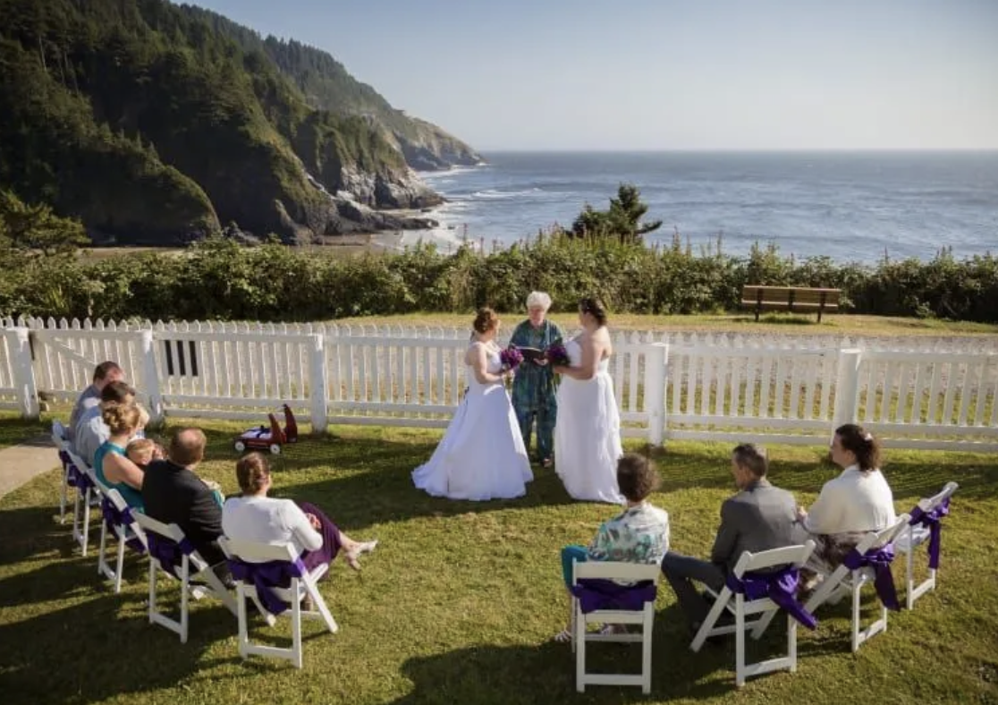 Wedding ceremony outdoors overlooking the ocean with a white picket fence, officiant, two brides in white dresses, and guests seated on white chairs with purple ribbons.