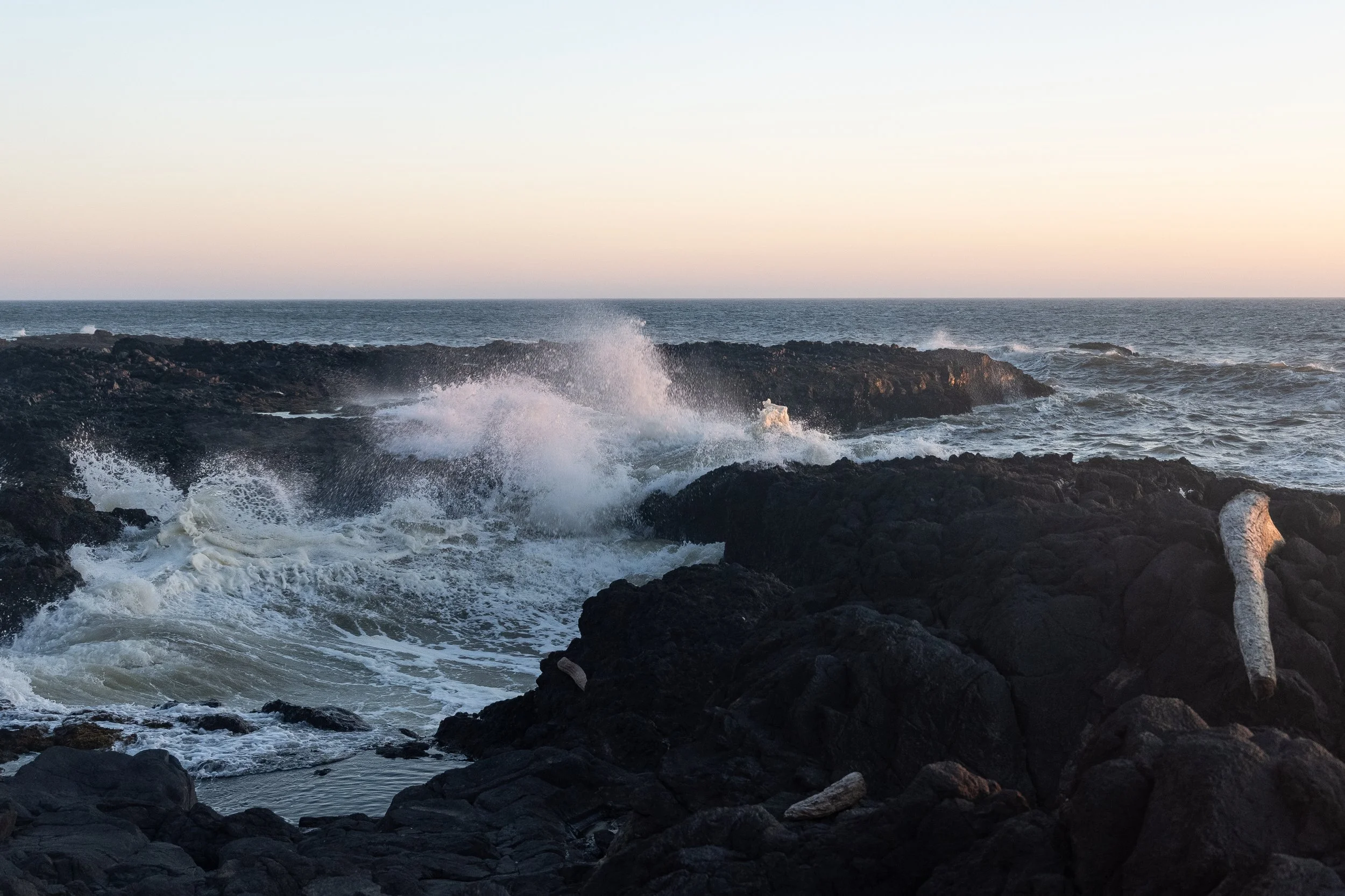 Waves crashing against dark volcanic rocks on a rocky shoreline at sunset with clear skies in the background.