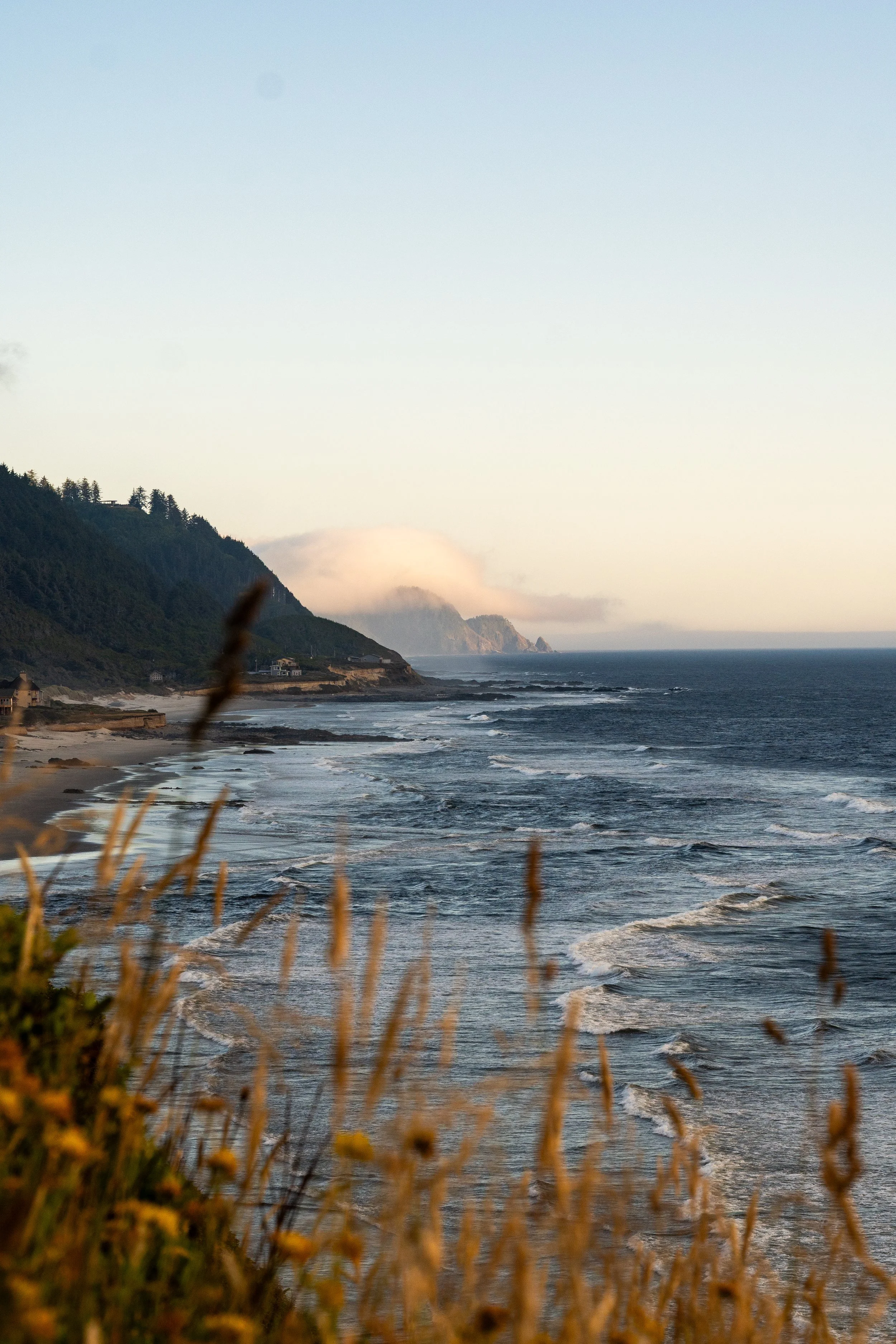 A scenic view of a beach with rolling waves, green hills in the background, and a partly cloudy sky during sunset or sunrise.