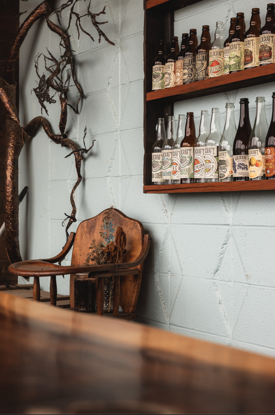 Wooden chair with floral design, dried flowers on it, next to a textured concrete wall with a branch decorating the left side, and a shelf above with various craft cider bottles.