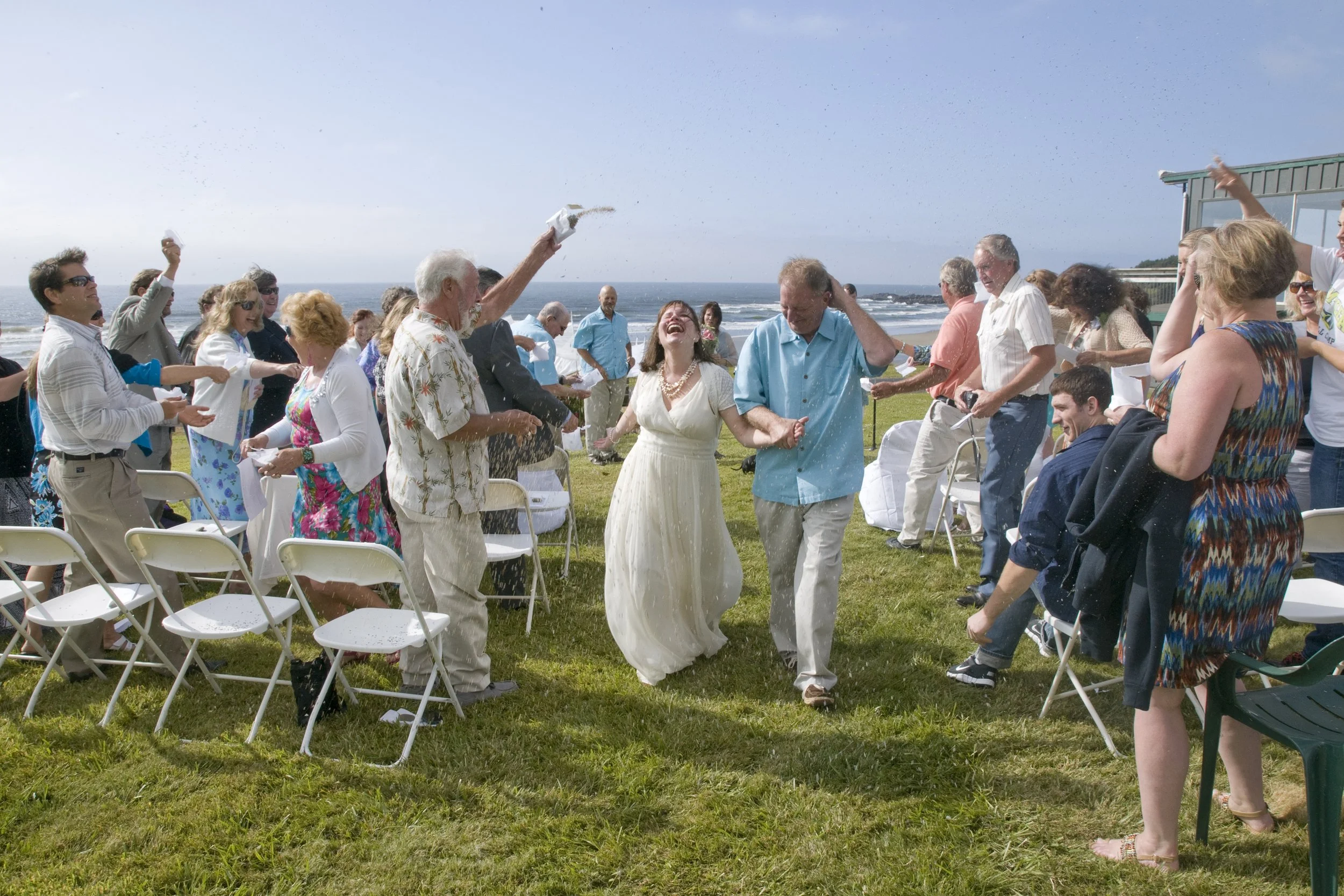 People celebrating a wedding outdoors at a beach, with the bride and groom dancing in the center while guests throw rice or confetti.