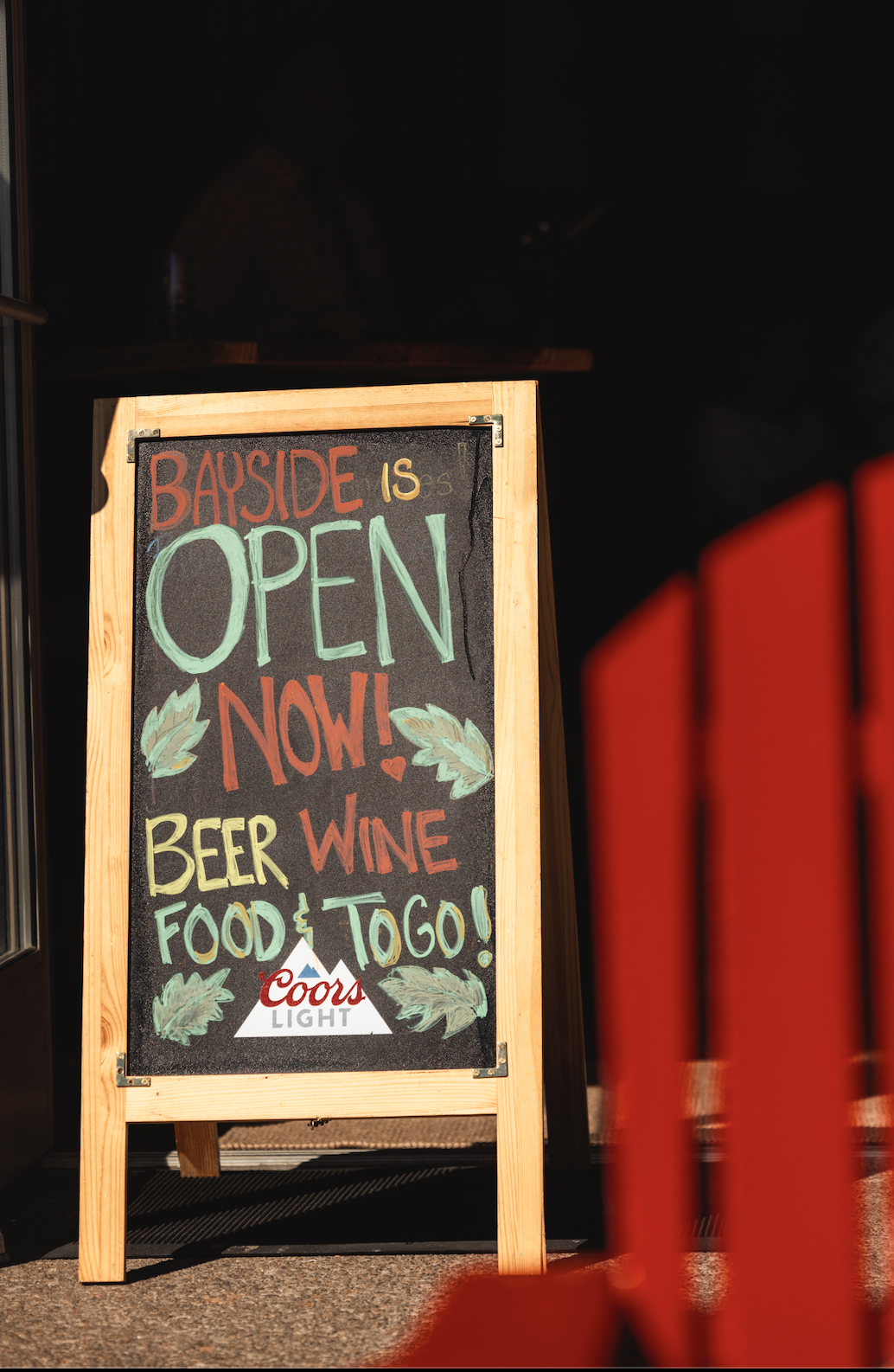 A chalkboard sign outside a bar or restaurant with colorful handwritten text reading "Bayside is open now! Beer, wine, food & to go!" and a Coors Light logo.