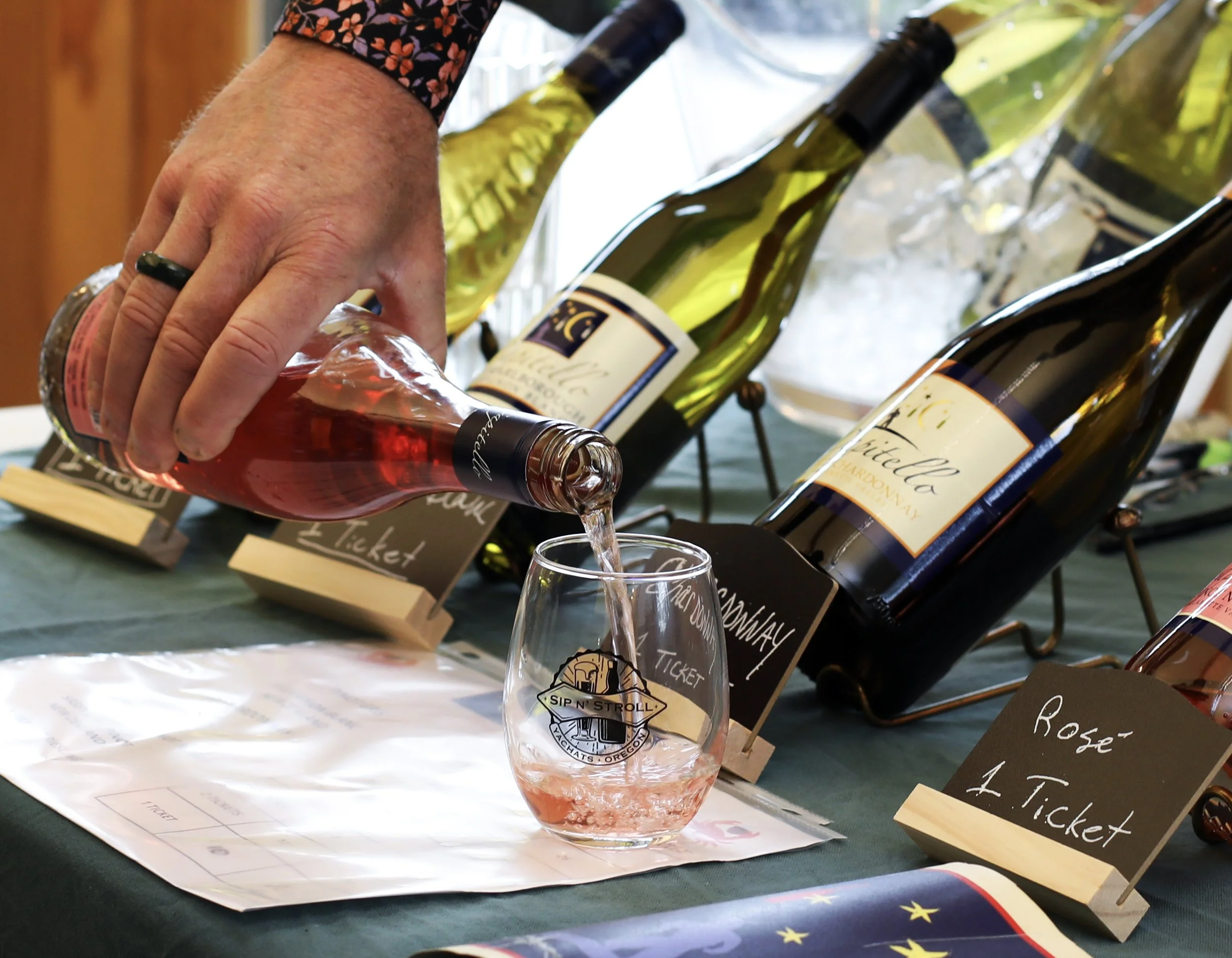 A person is pouring pink wine from a bottle into a small glass with a logo on it. There are several bottles of white and rosé wine displayed on a table with black signs indicating prices and types of wine. The setting appears to be a wine tasting eve
