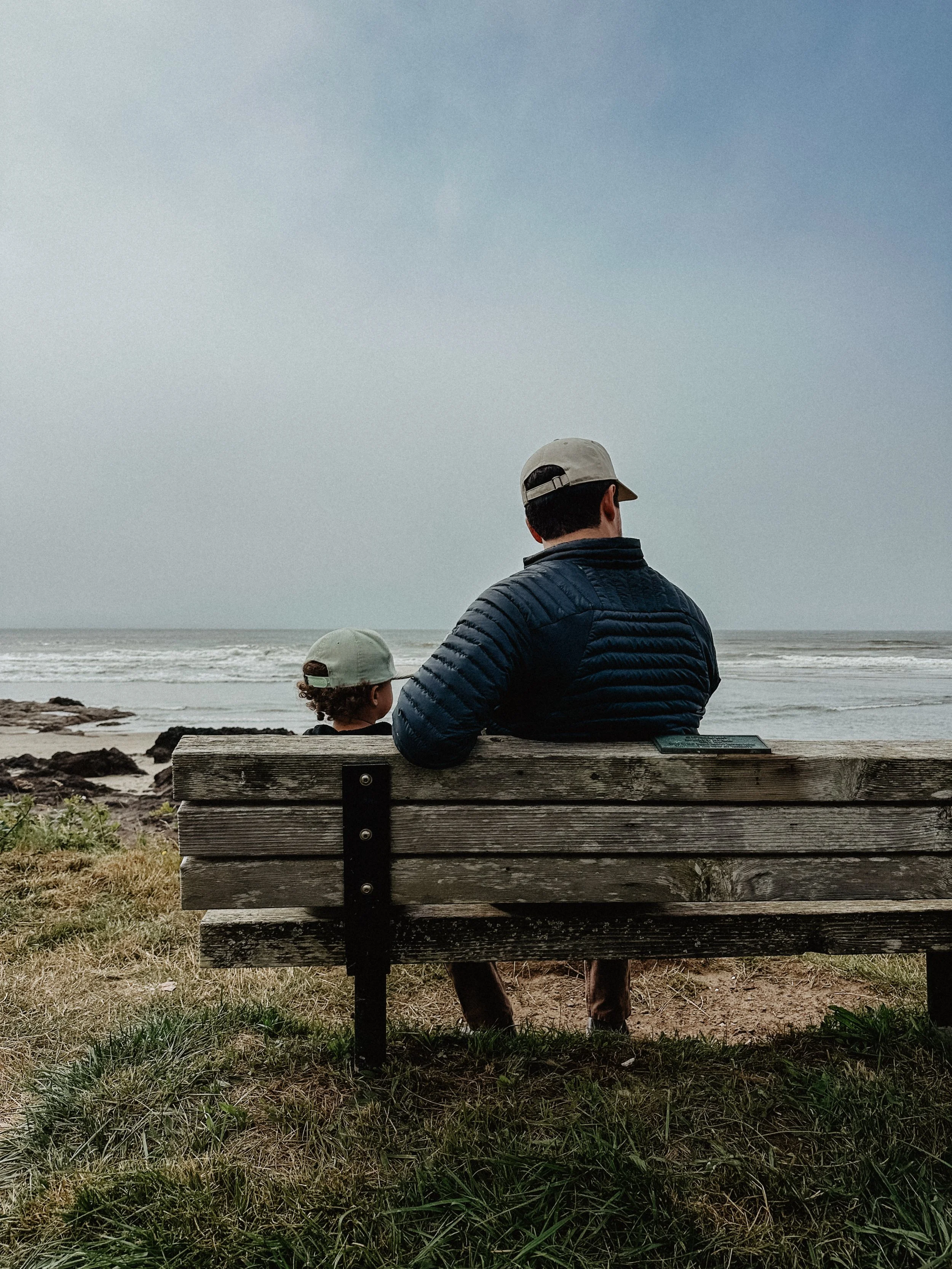 Man and child sitting on a wooden bench, facing the ocean on a cloudy day.