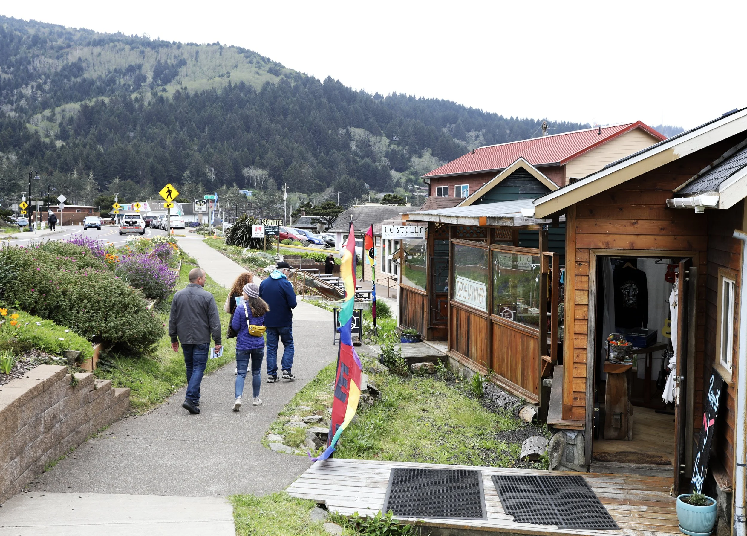 People walking along a sidewalk in a small mountain town, with colorful flags and wooden storefronts, surrounded by green hills and flowering bushes.