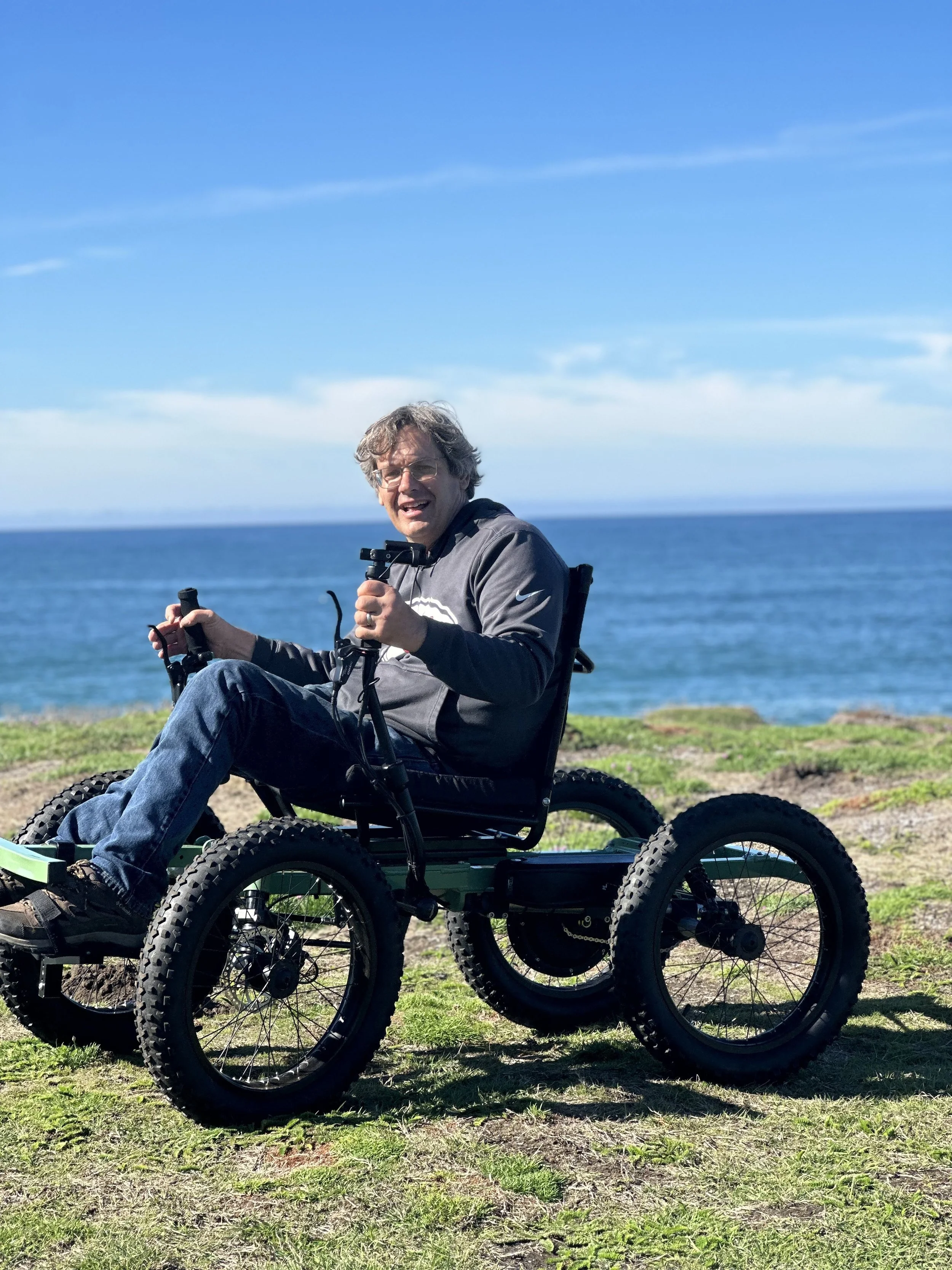 Man in a gray hoodie on a motorized off-road wheelchair near the ocean on a sunny day.