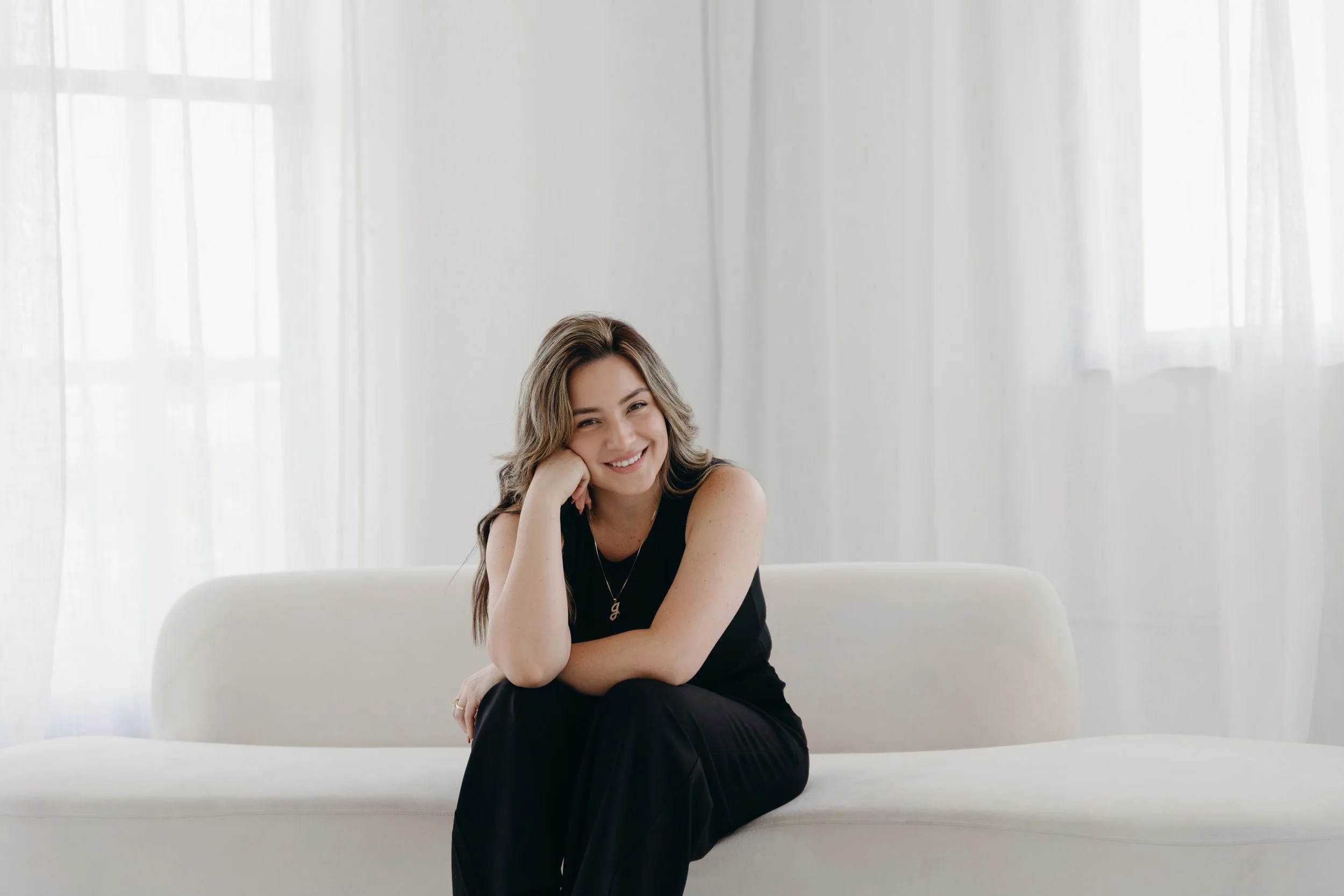 A smiling woman with wavy hair, wearing a black sleeveless top, sitting on a white sofa in a bright room with sheer white curtains and large windows.