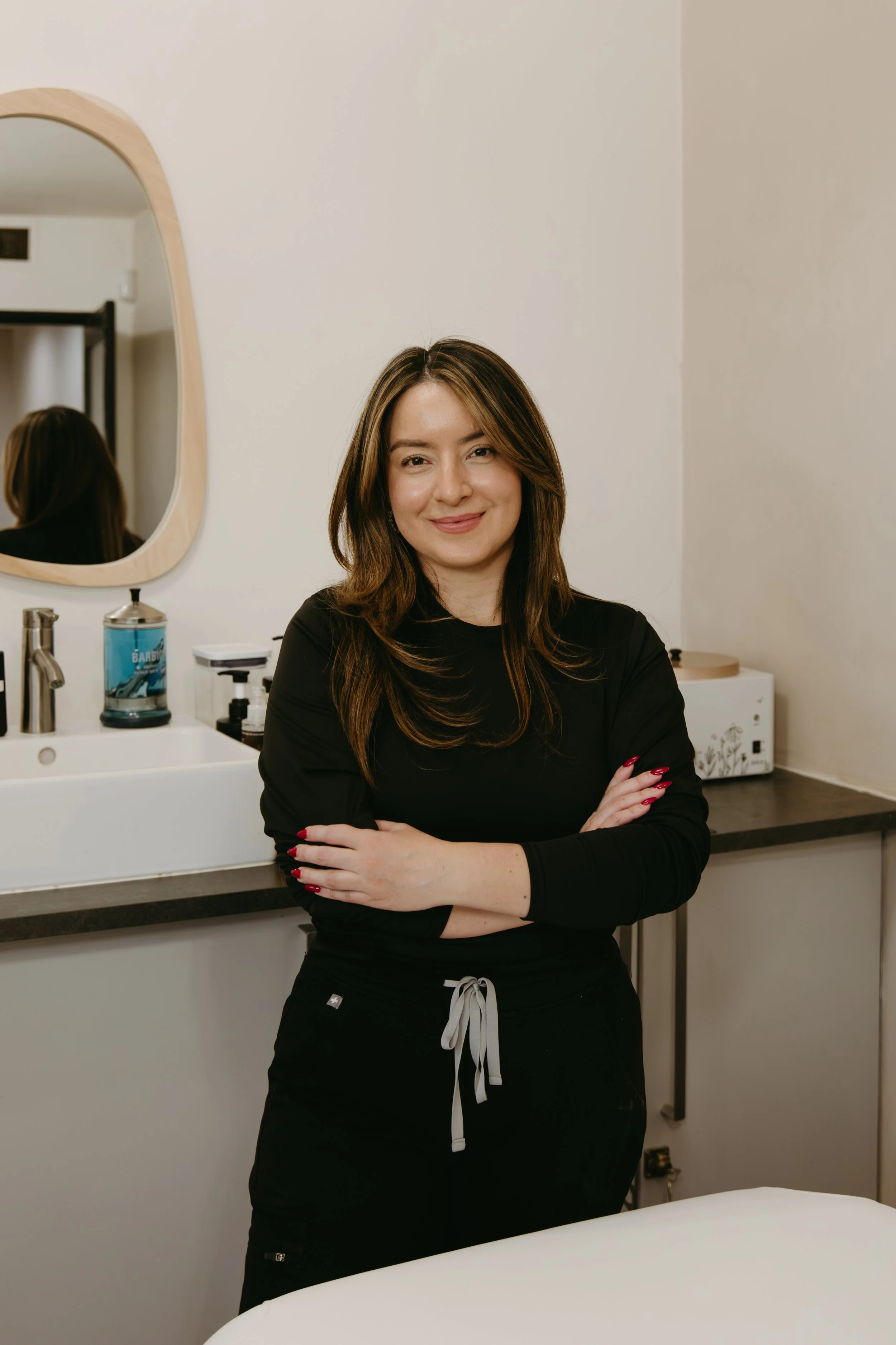 A woman with light skin and brown hair smiling and crossing her arms, standing in a room with a mirror, sink, and countertop.