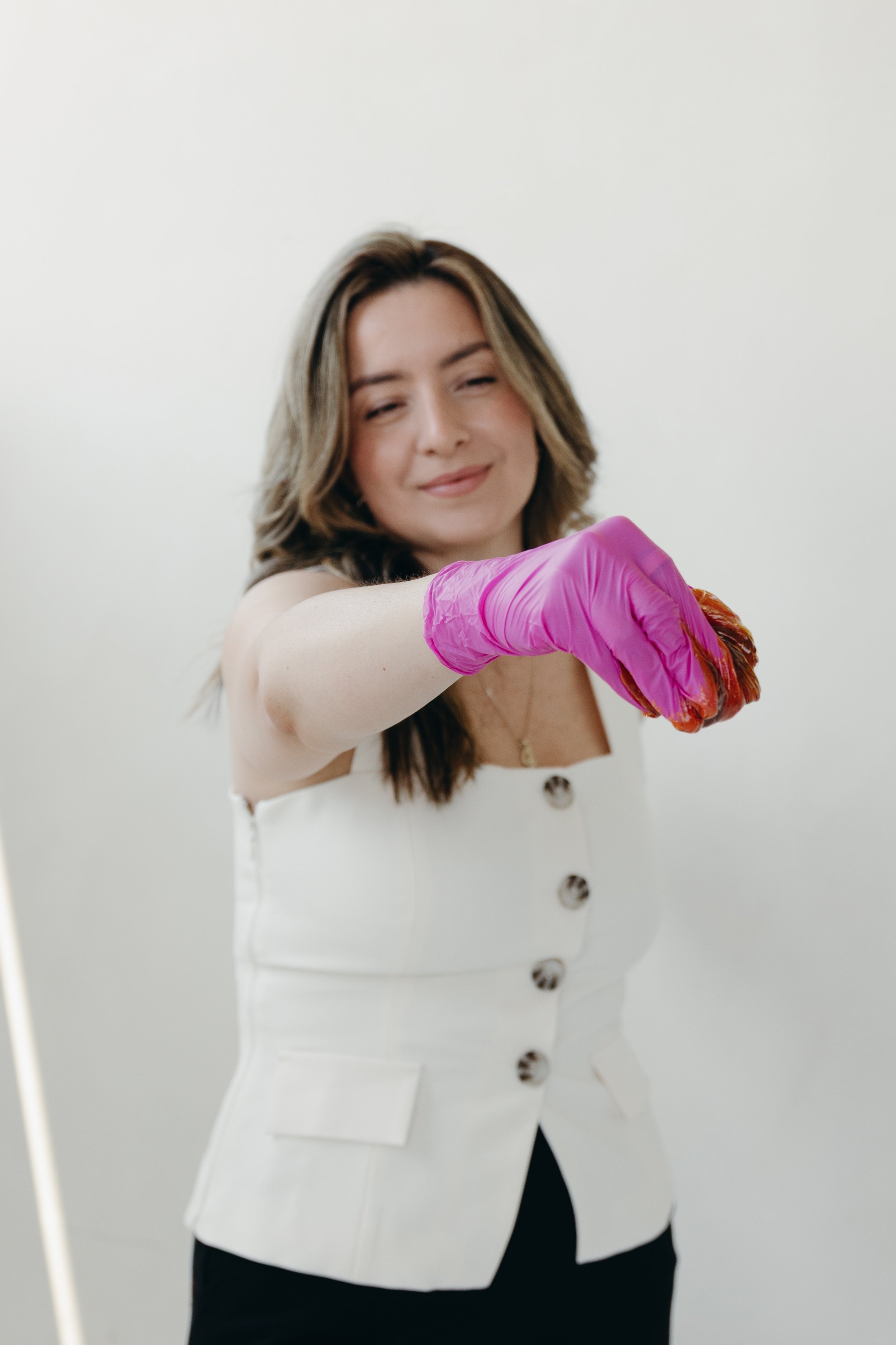 Woman with brown hair wearing a white sleeveless blazer and pink gloves, holding a piece of meat.
