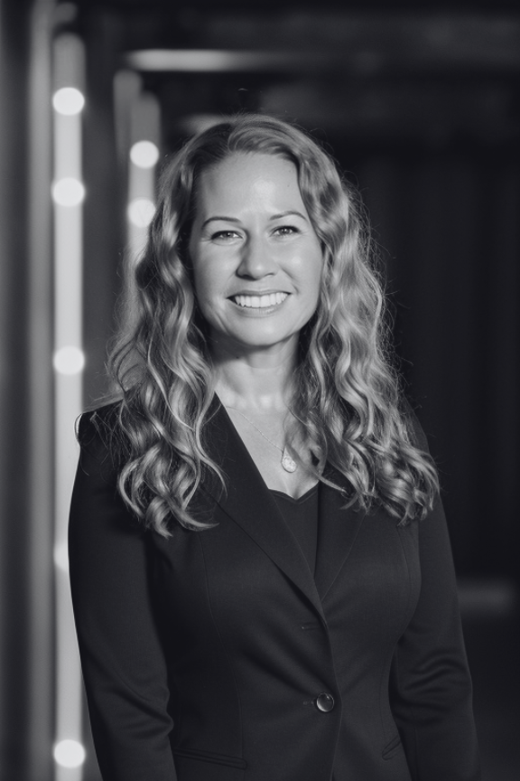 A smiling woman with long wavy hair, wearing a black blazer and a necklace, standing in a modern indoor setting with blurred lights in the background.