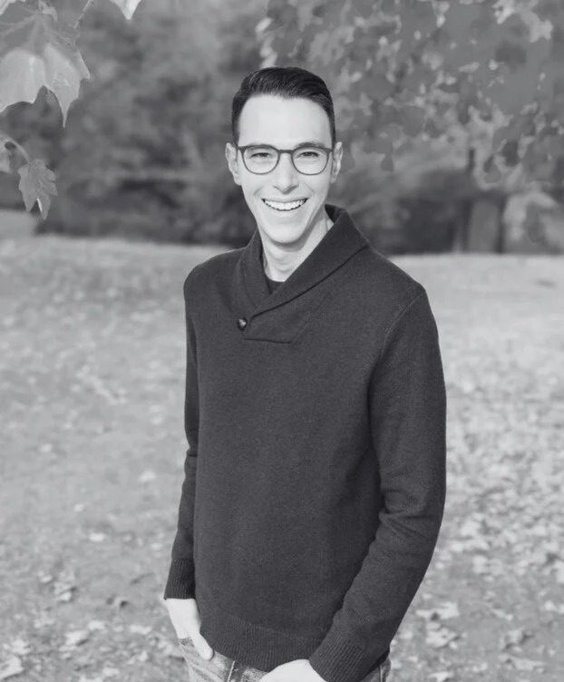 A smiling young man with glasses standing outdoors on a gravel path, surrounded by trees.