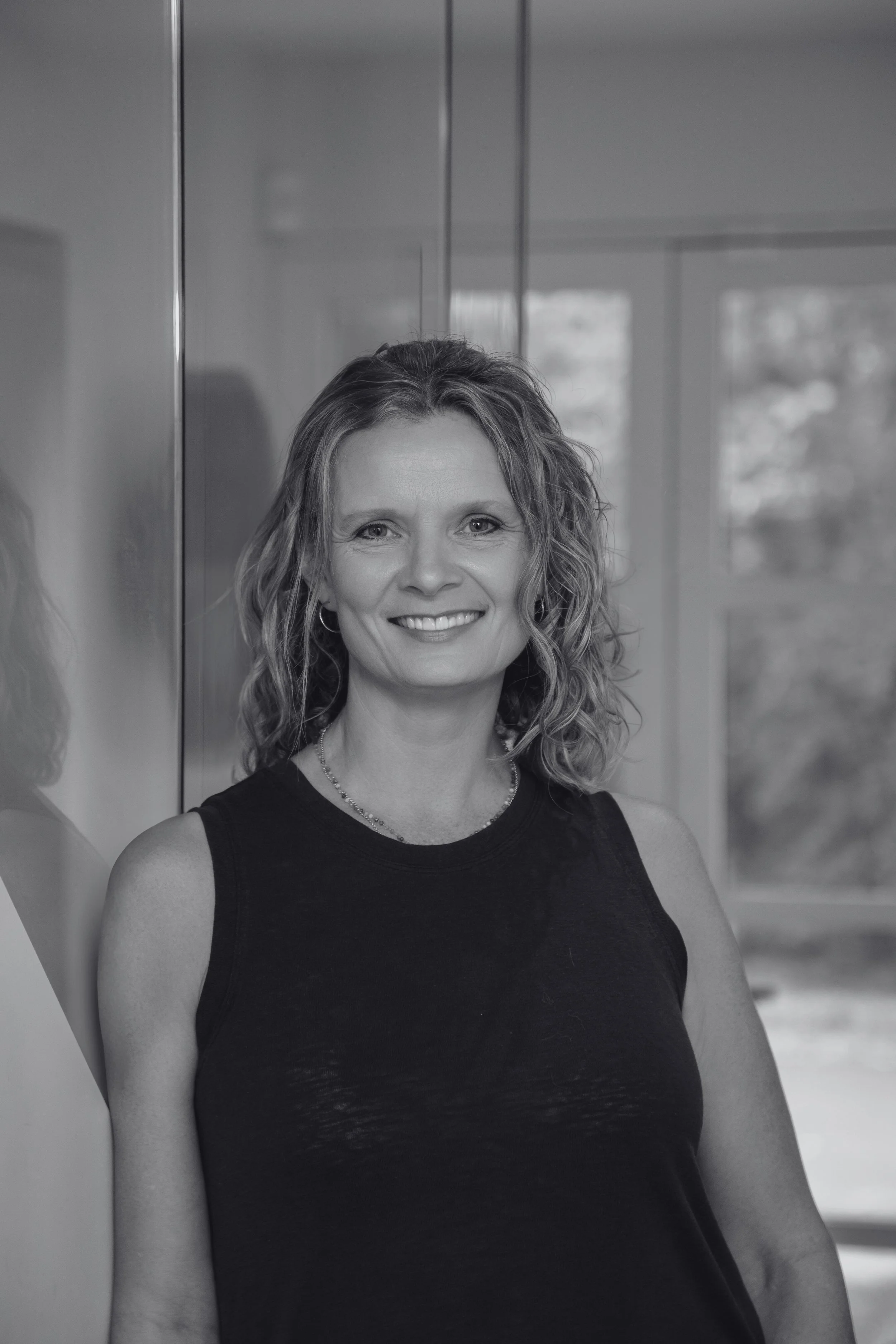 Black and white portrait of a smiling woman with wavy hair, wearing a sleeveless top, standing against a reflective surface with a window in the background.