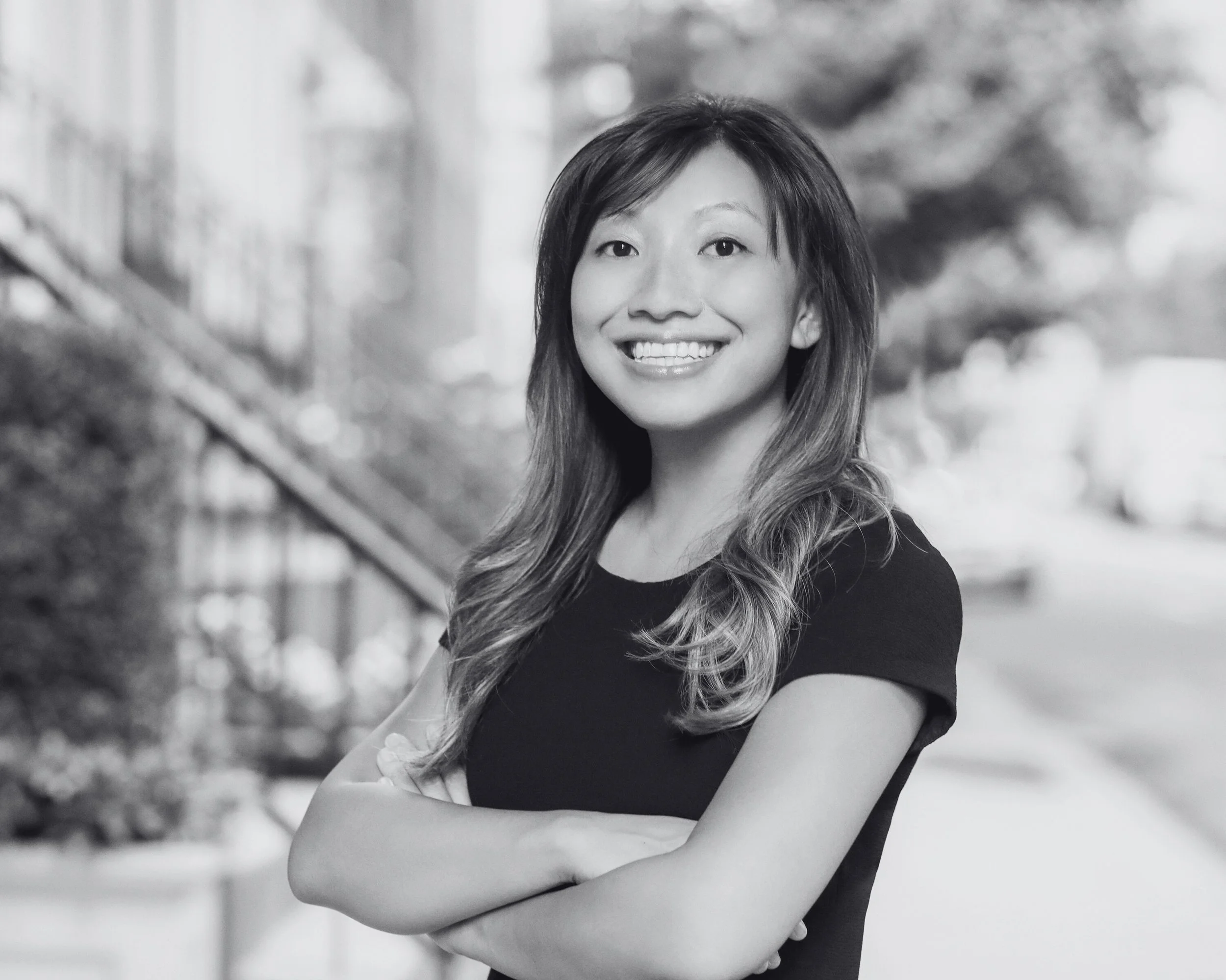 A smiling woman with long wavy hair standing outdoors with her arms crossed, background of a blurred street scene.